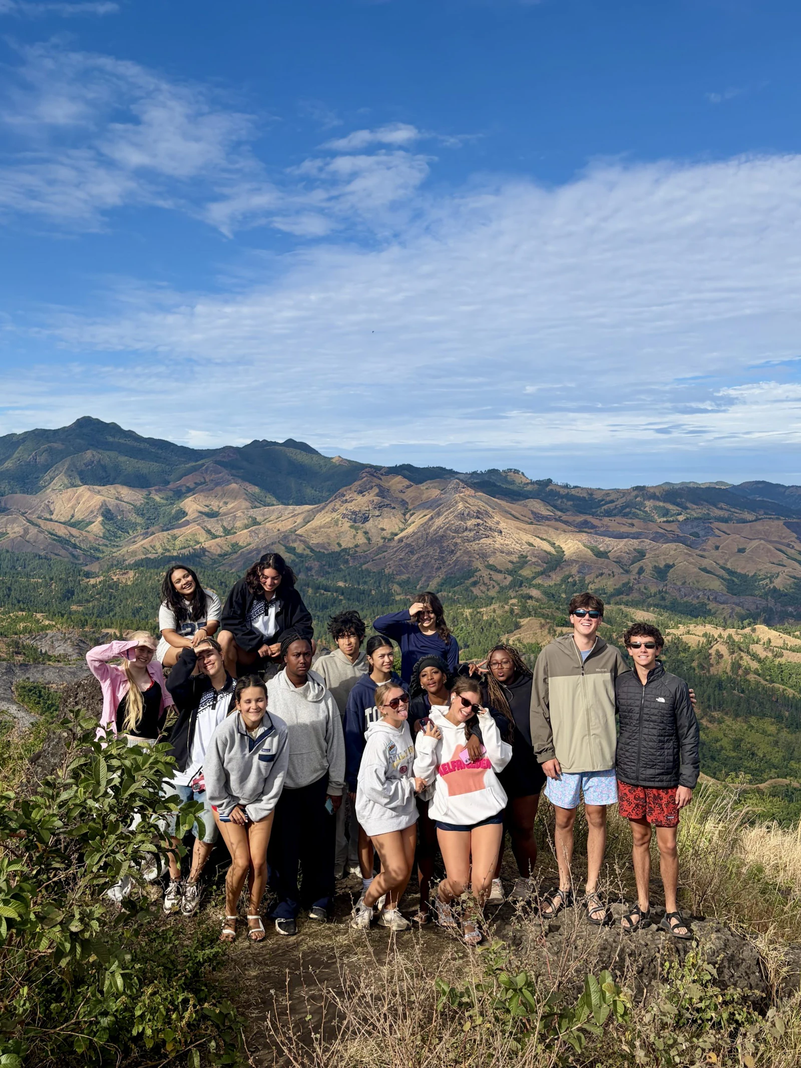 Teen travelers stand on a ridge in Fiji with red and green mountain ranges behind them