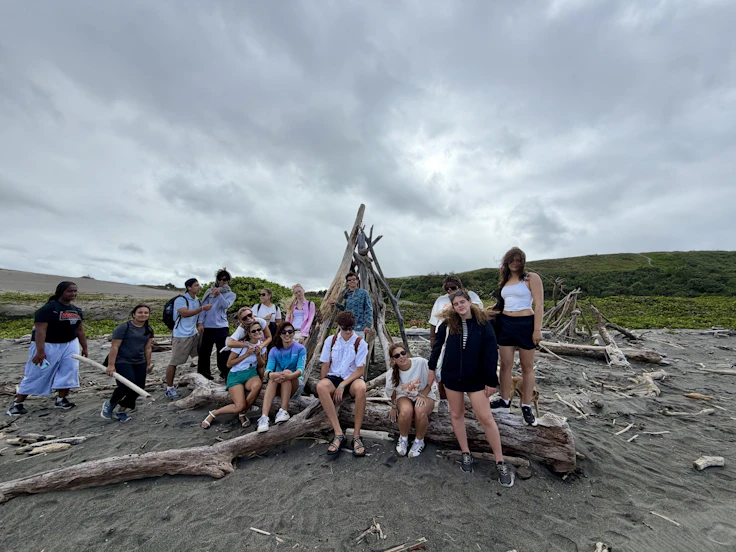 Teen travelers sit along bleached driftwood logs around a teepee frame on a grey Fijian beach under cloud.