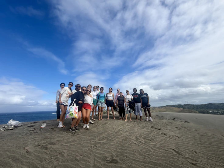 Teen travelers stand together on a wide grey sand dune above the sea in Fiji under a big cloudy sky.