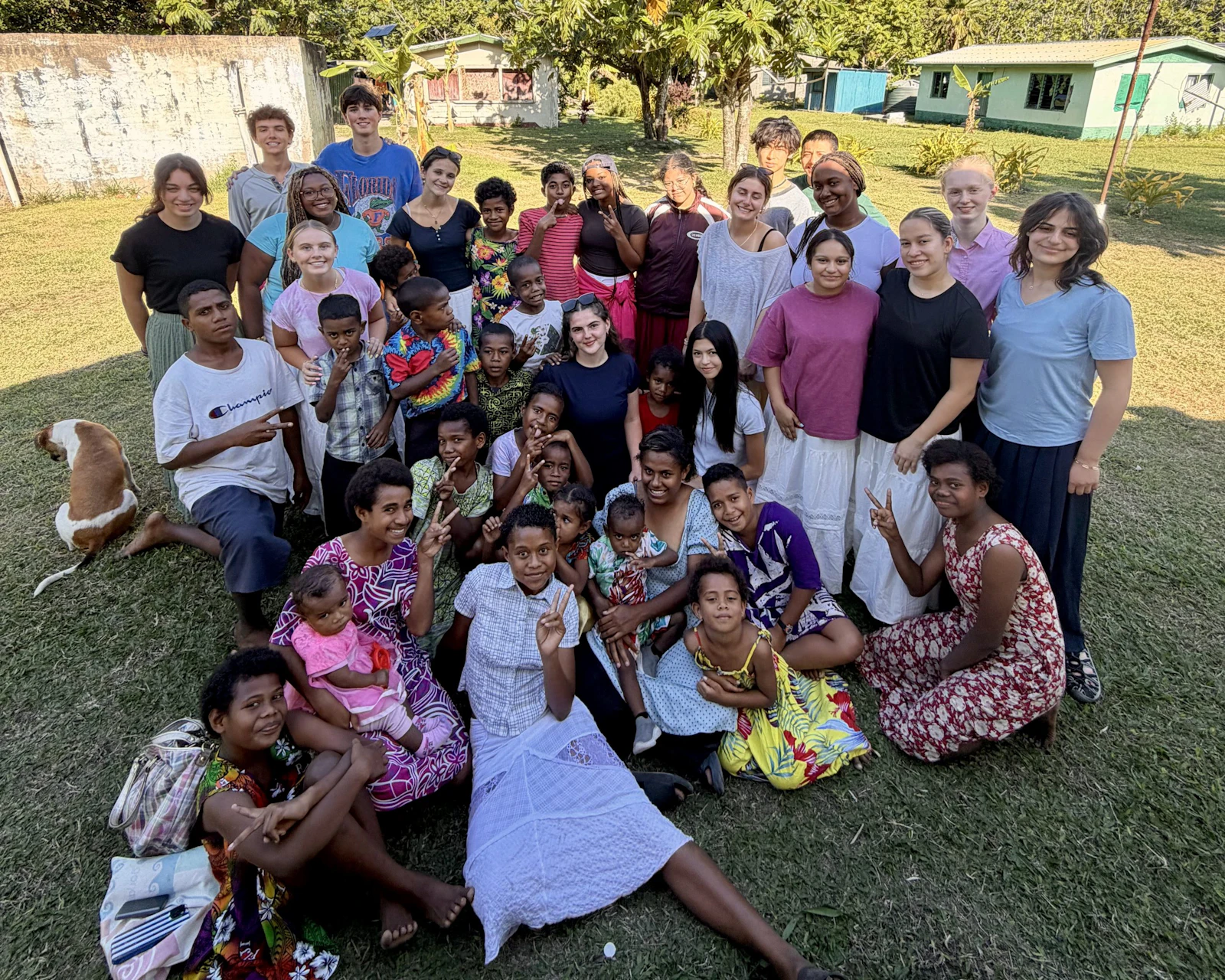 Teen travelers sit and stand on the grass with Fijian women and children outside village houses