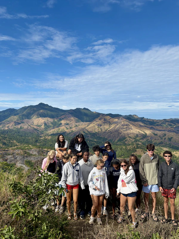 The full group of teen travelers stands on a Fijian ridge with eroded green ranges spread out behind them