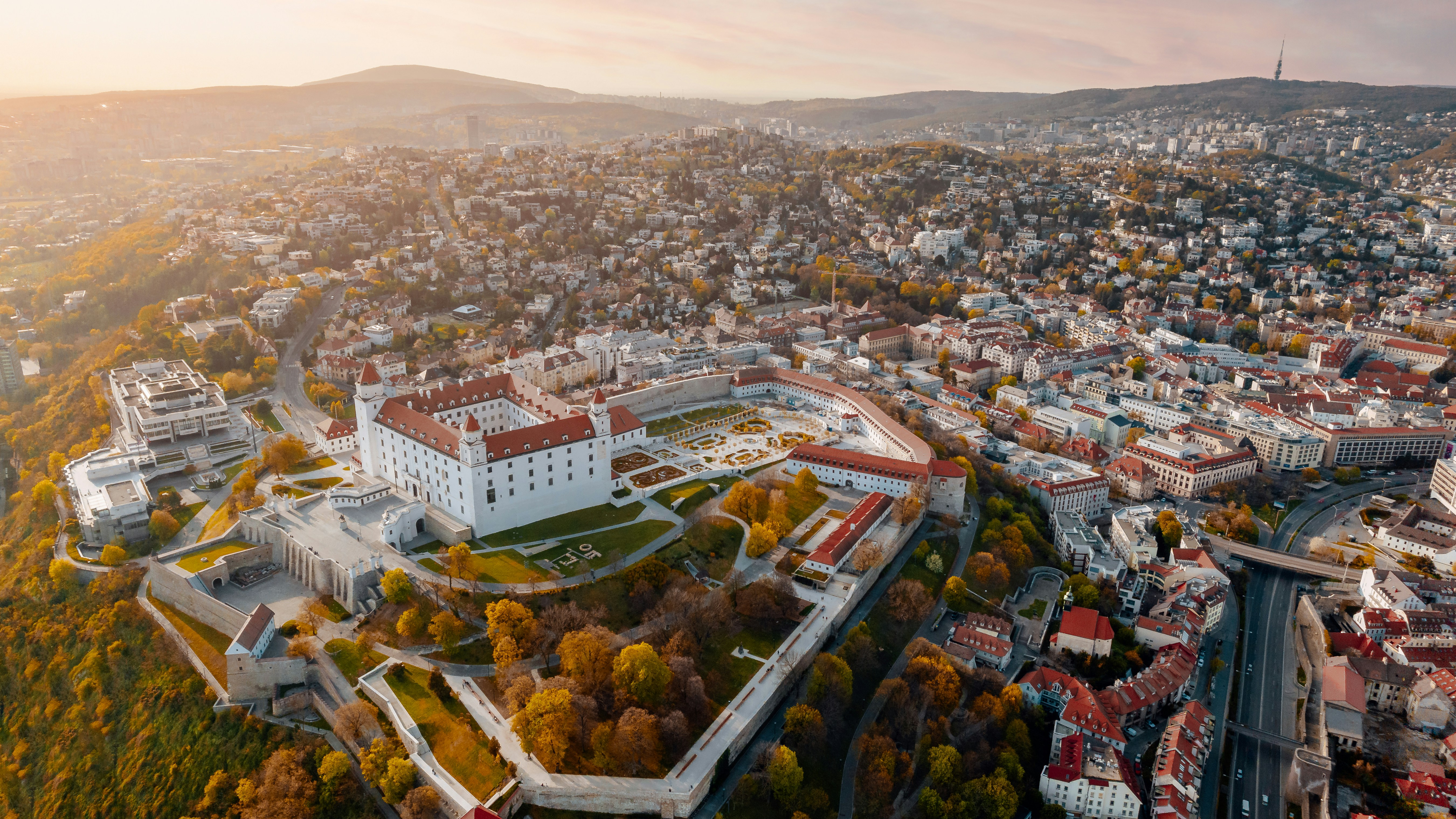 An aerial view of Bratislava Castle above the city's rooftops and autumn trees at sunrise.
