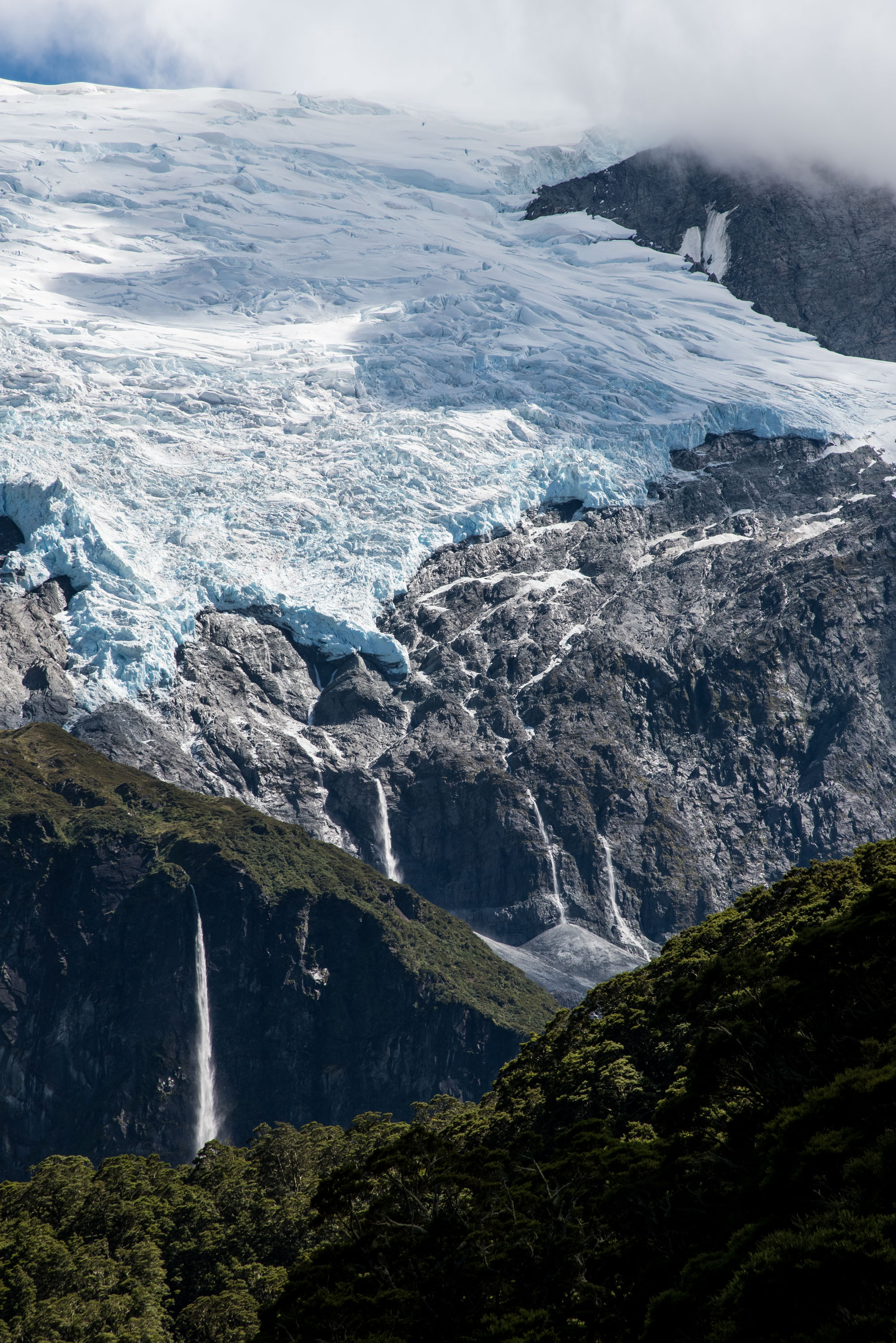 A glacier spills down a rocky mountainside above waterfalls and forest