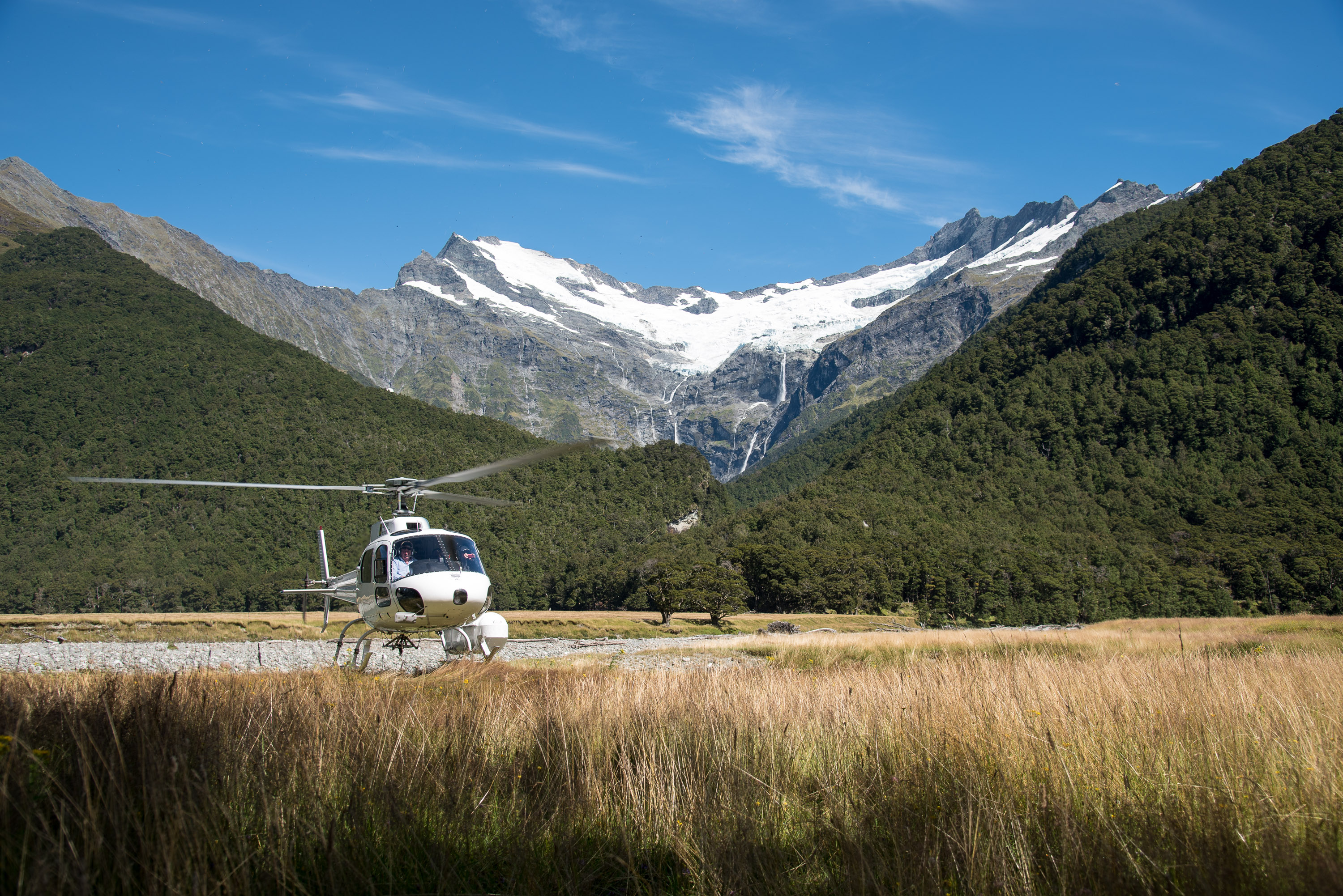 A helicopter waits in a grassy valley beneath snow-capped mountains