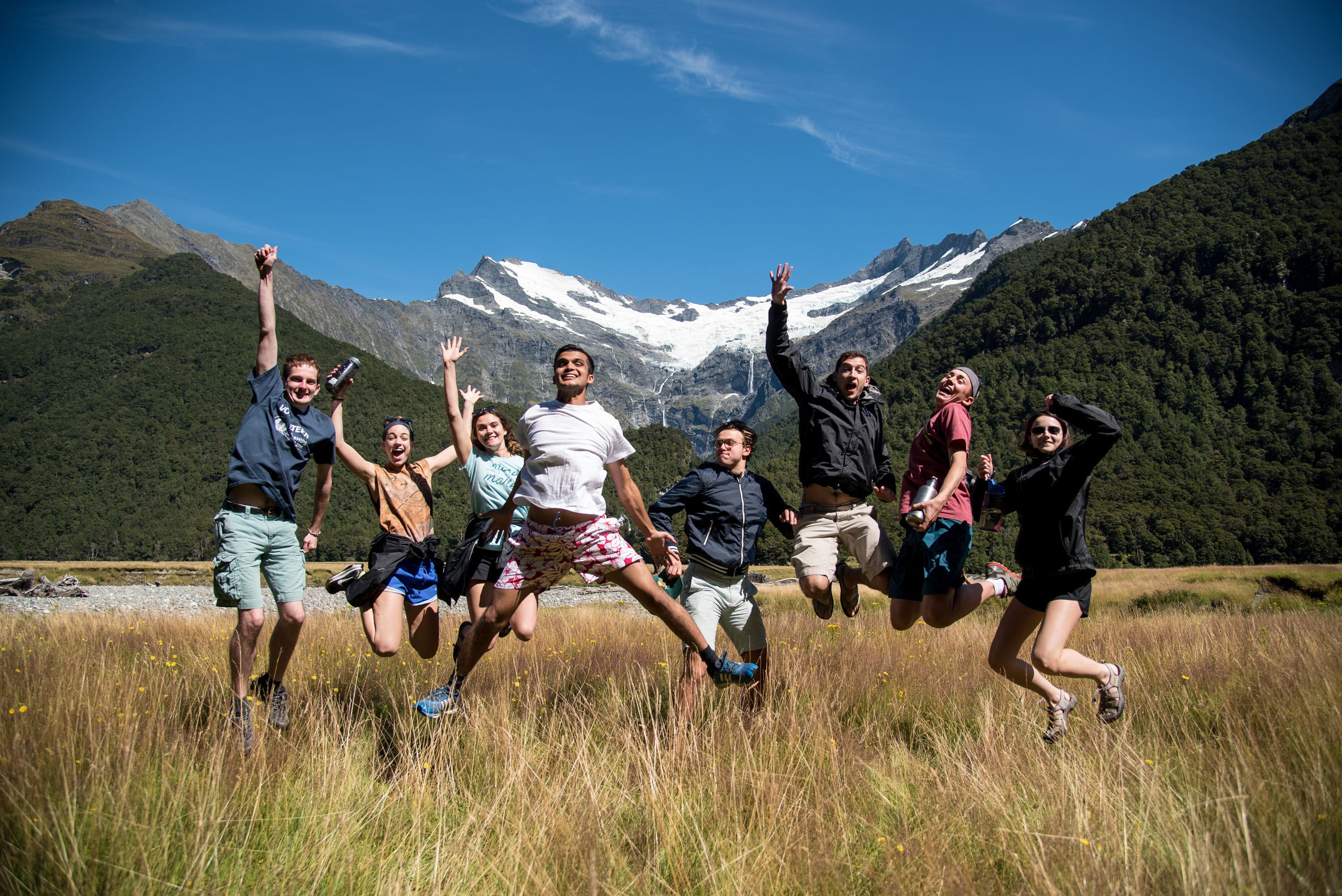 Students jump for joy in a grassy valley beneath snow-capped mountains