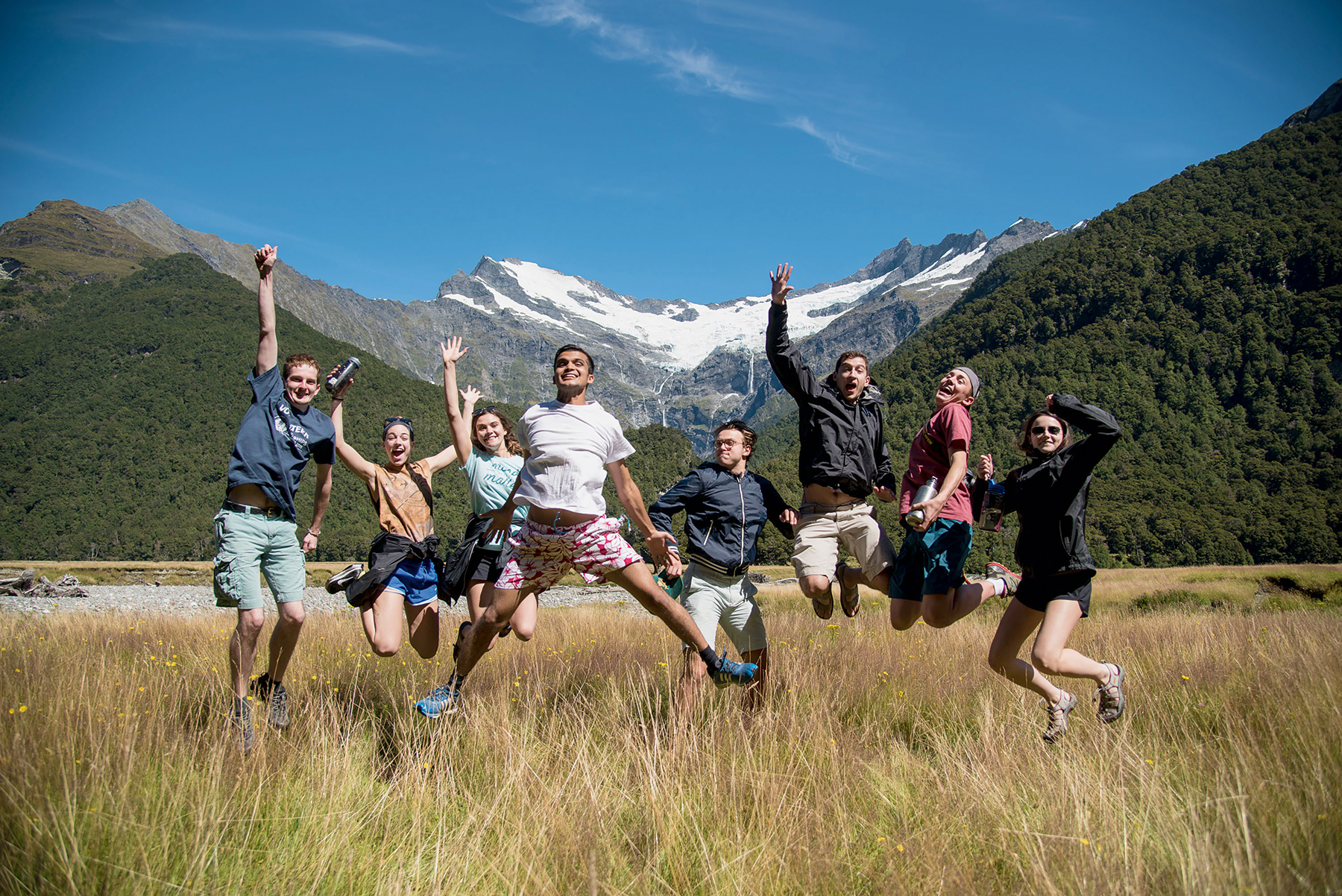 Students jump for joy in a grassy valley below snow-capped mountains