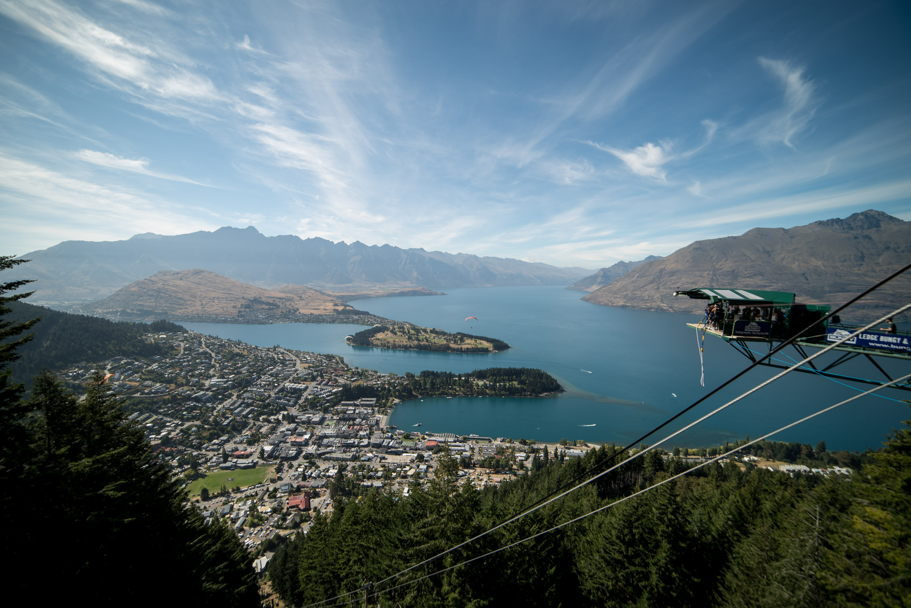 A lake town on a green shoreline seen from high above, with a long jagged snow-streaked range across the water.