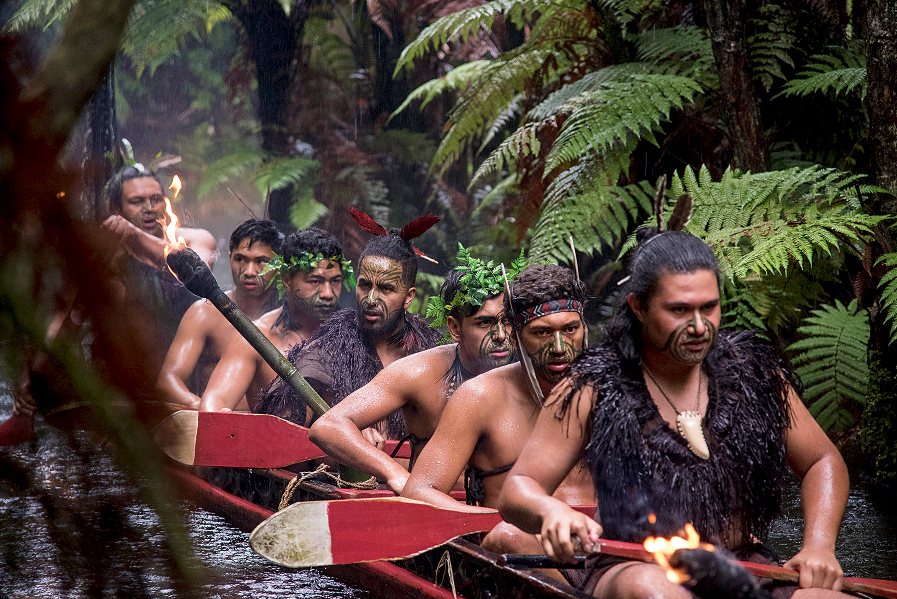 Performers in traditional dress paddle a canoe through a fern-lined waterway