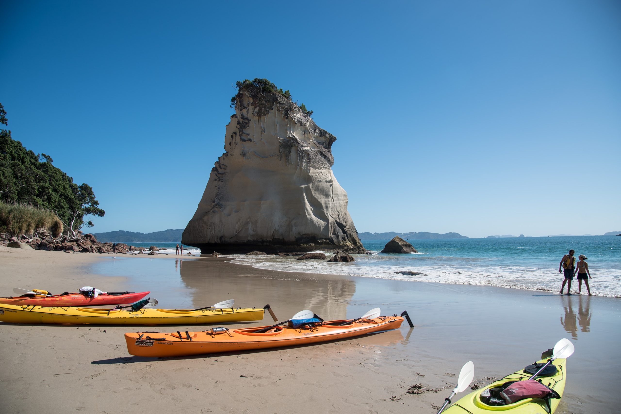 Sea kayaks pulled up on a sandy beach in front of a tall rock stack, with two people at the water's edge.