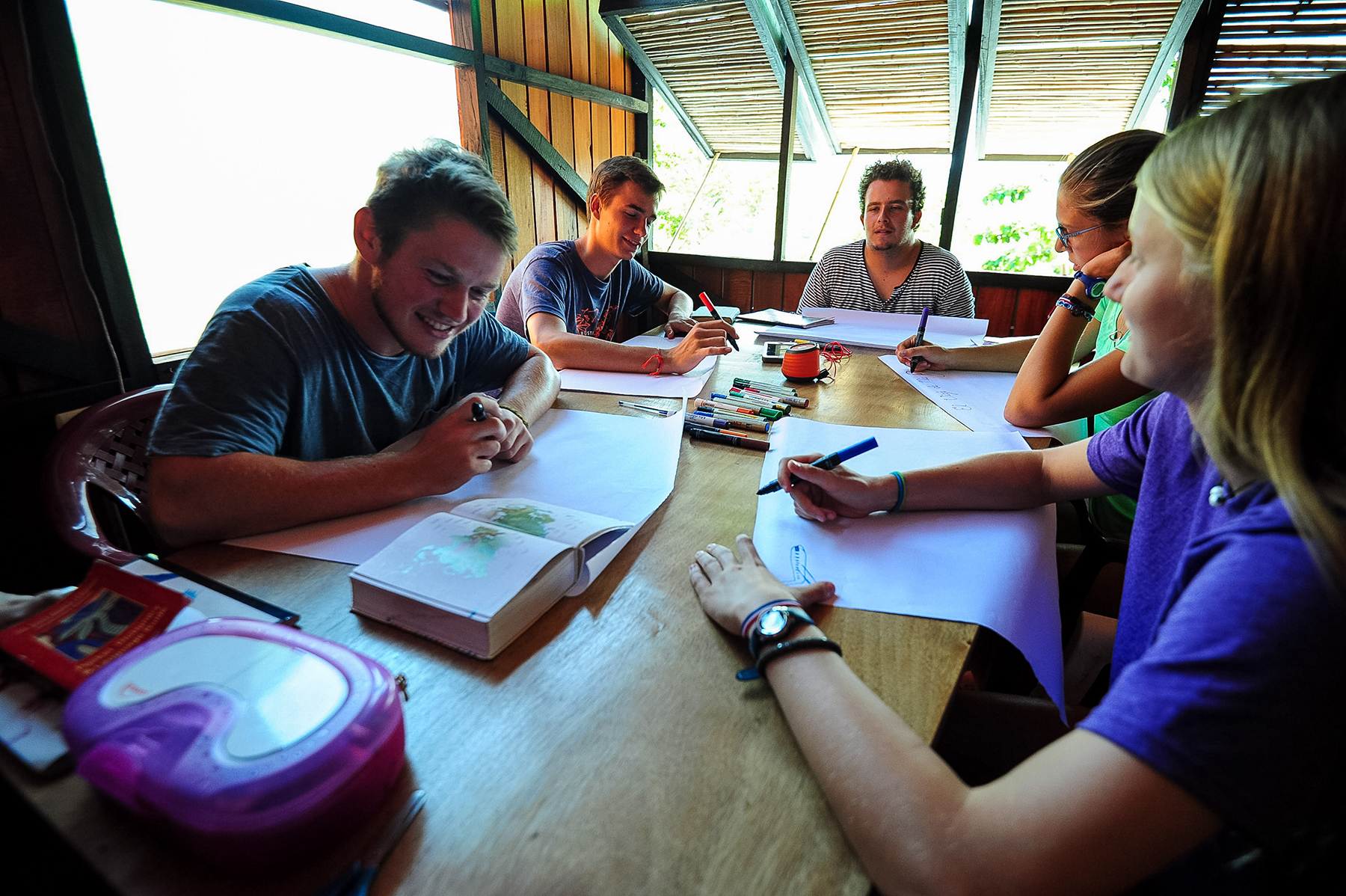 Students sit around a wooden table writing in notebooks under an open-air roof