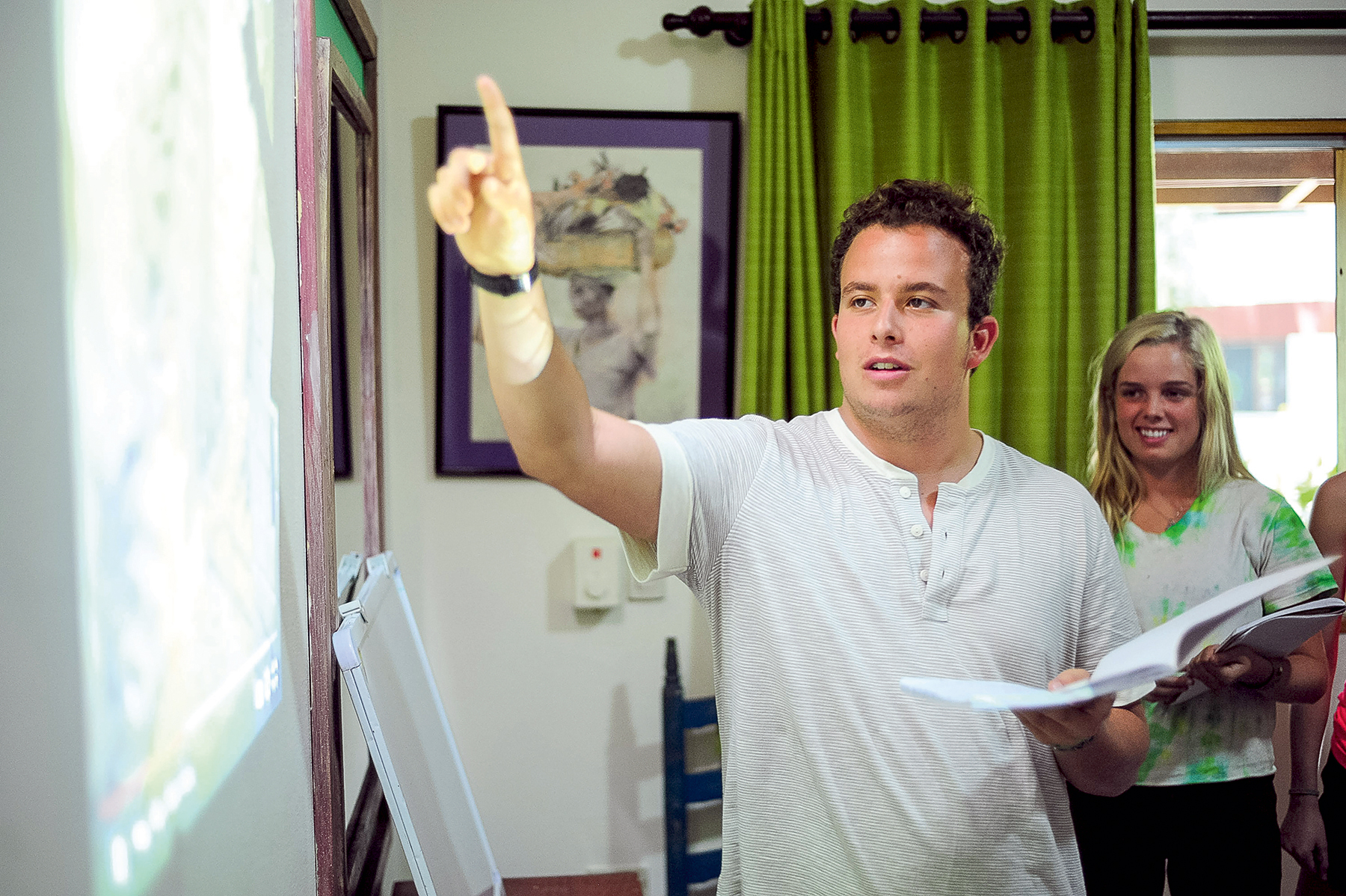 An instructor points at a projector screen while student travelers hold notebooks nearby