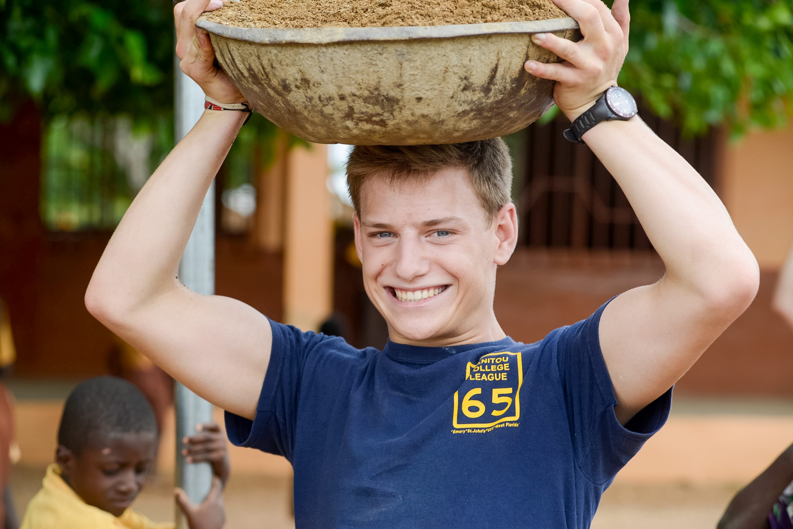 A student in Ghana smiles while balancing a large bowl of sand on his head