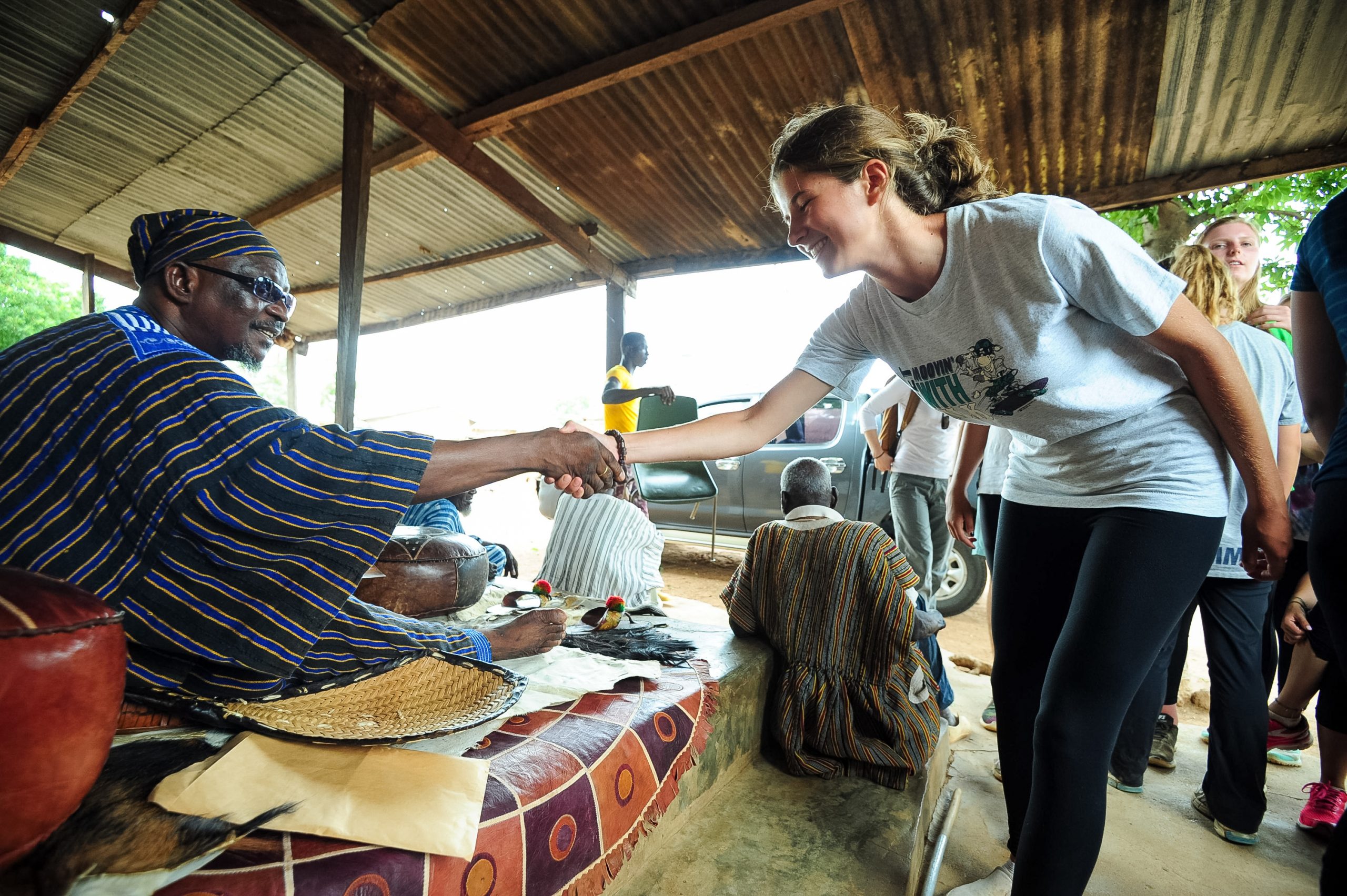 A student shakes hands with a seated community elder under a thatched shelter