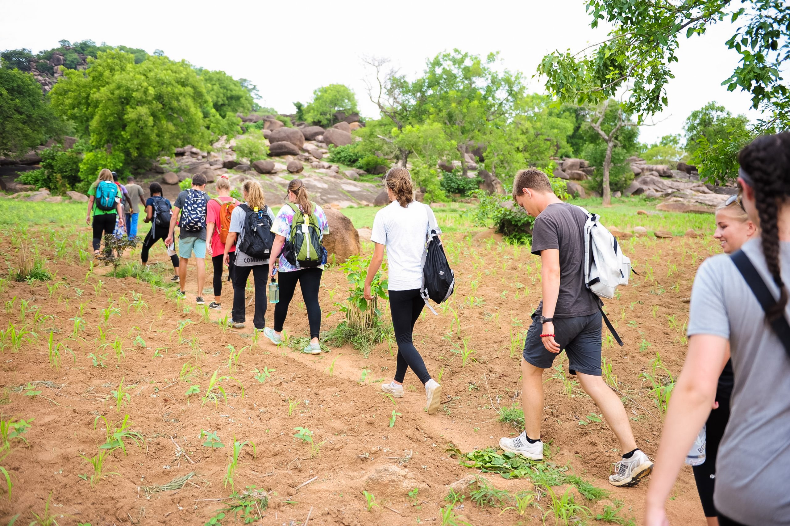 A school trip winds single file along a dirt path through rocky, green terrain in Ghana