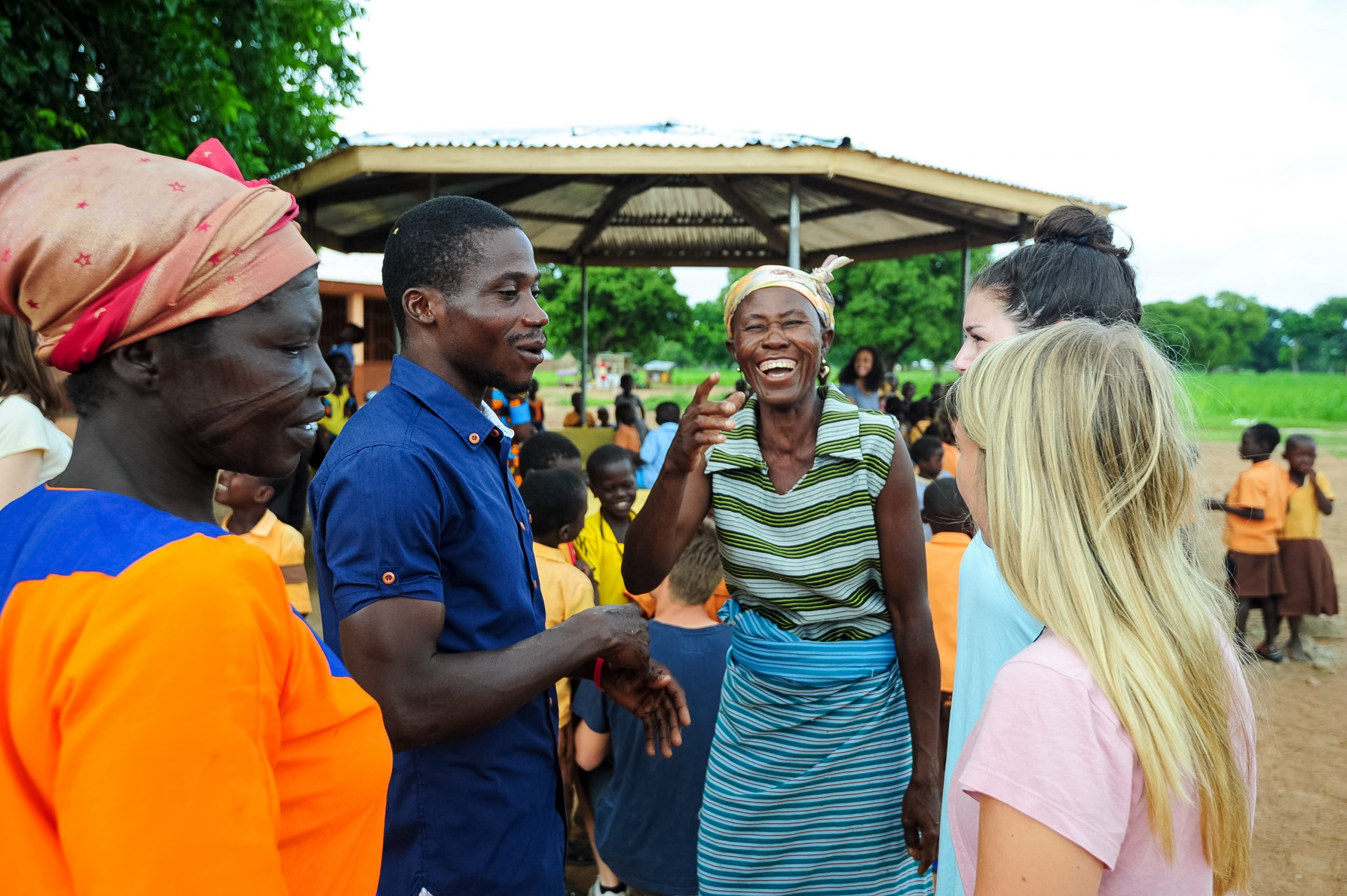 Students share a laugh with local community members outdoors under a thatched shelter