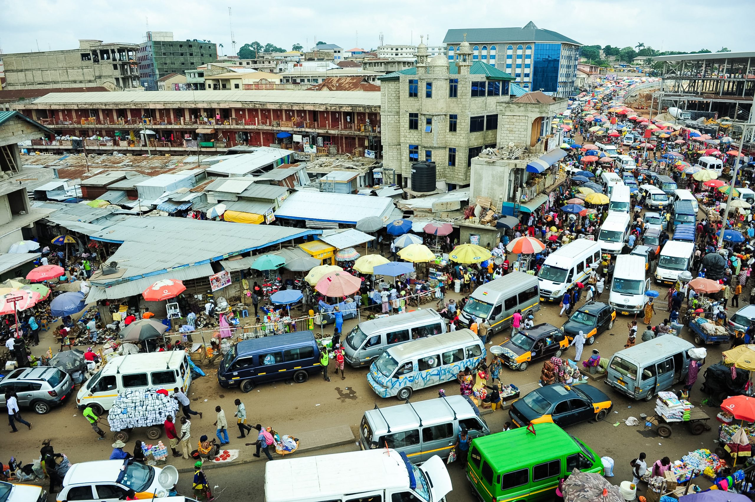 An aerial view overlooks a crowded market street lined with colorful umbrellas, vans, and vendors