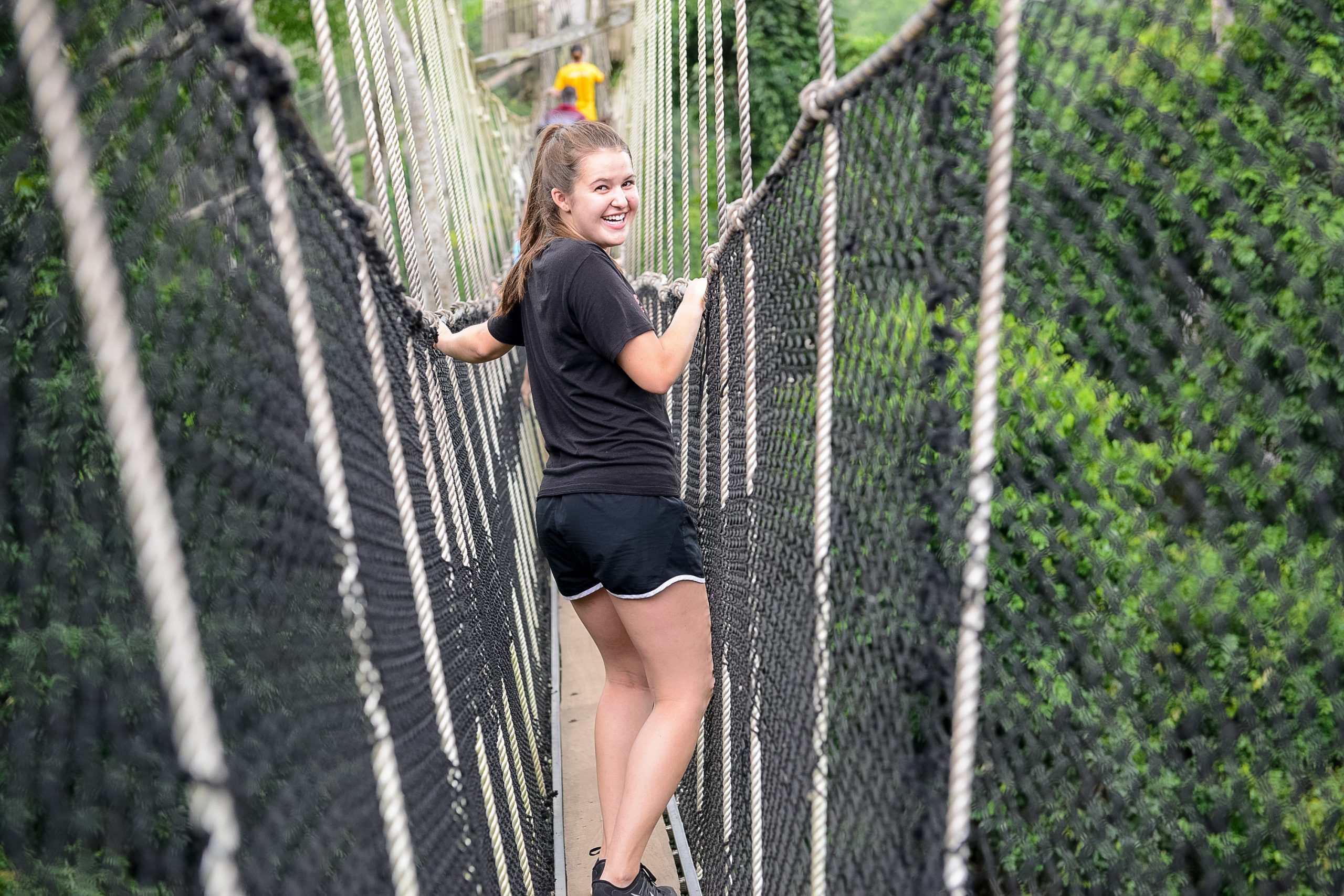 A student smiles back while crossing a rope canopy bridge in the rainforest