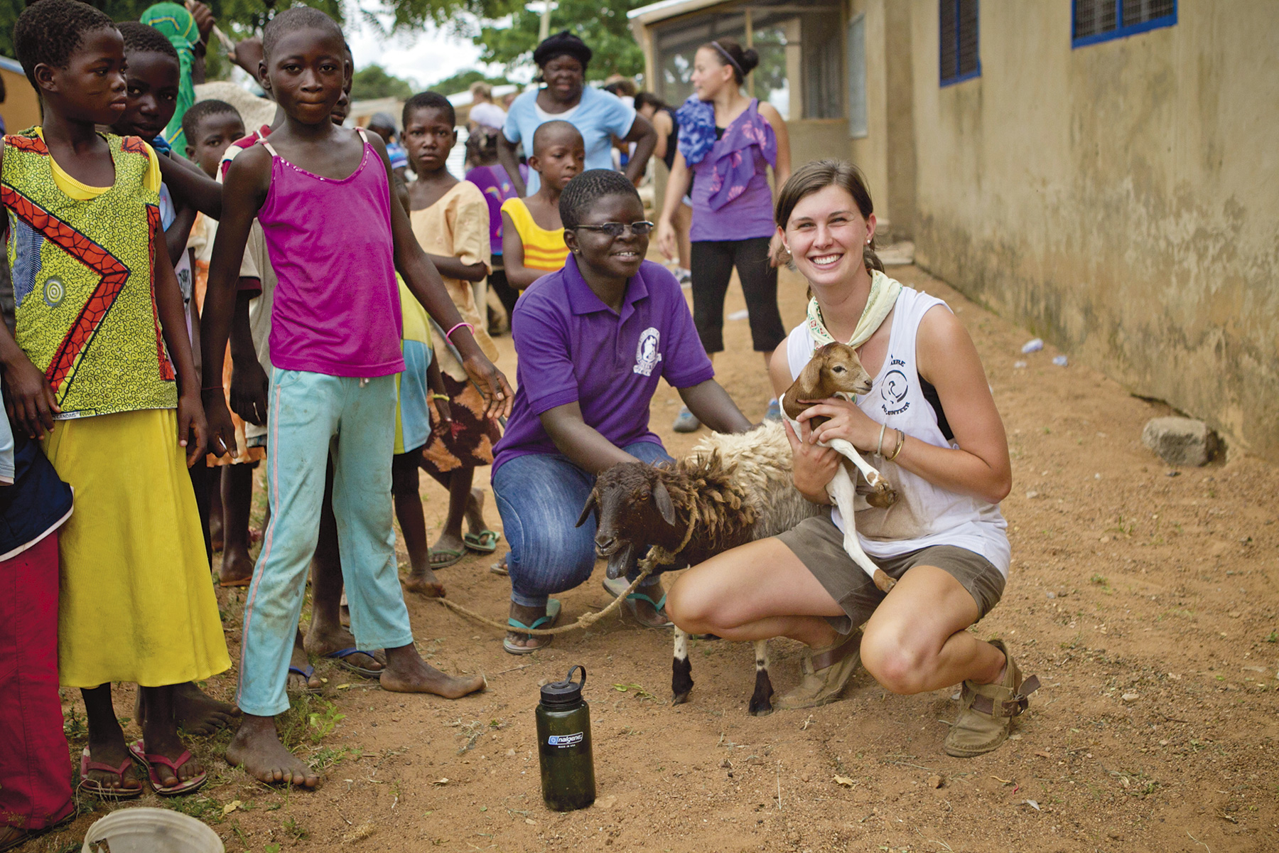 A student and a local leader crouch with a young goat as children gather around in a village courtyard