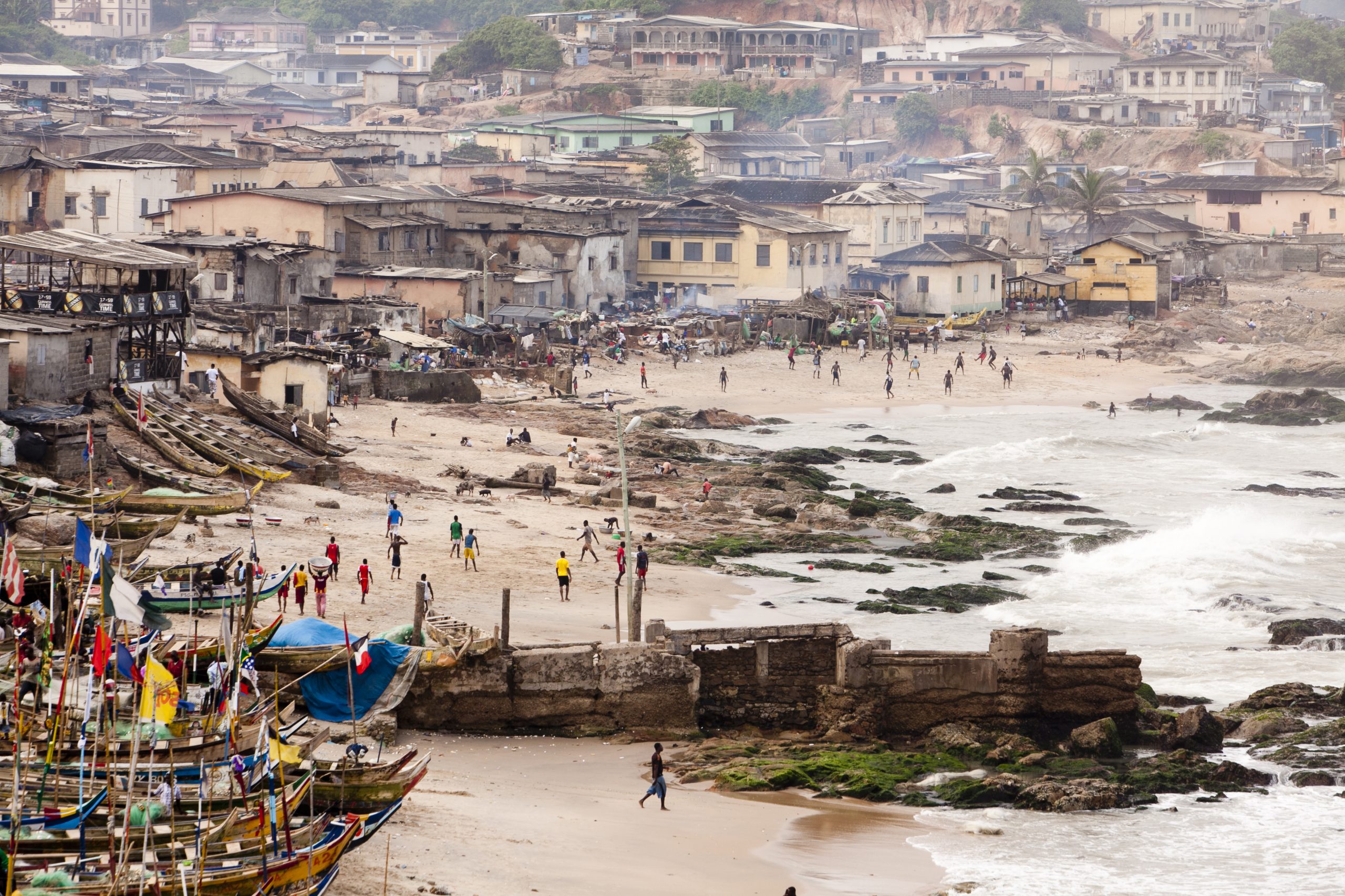 Fishing boats line a beach below a coastal town, with waves rolling in past a stone breakwater