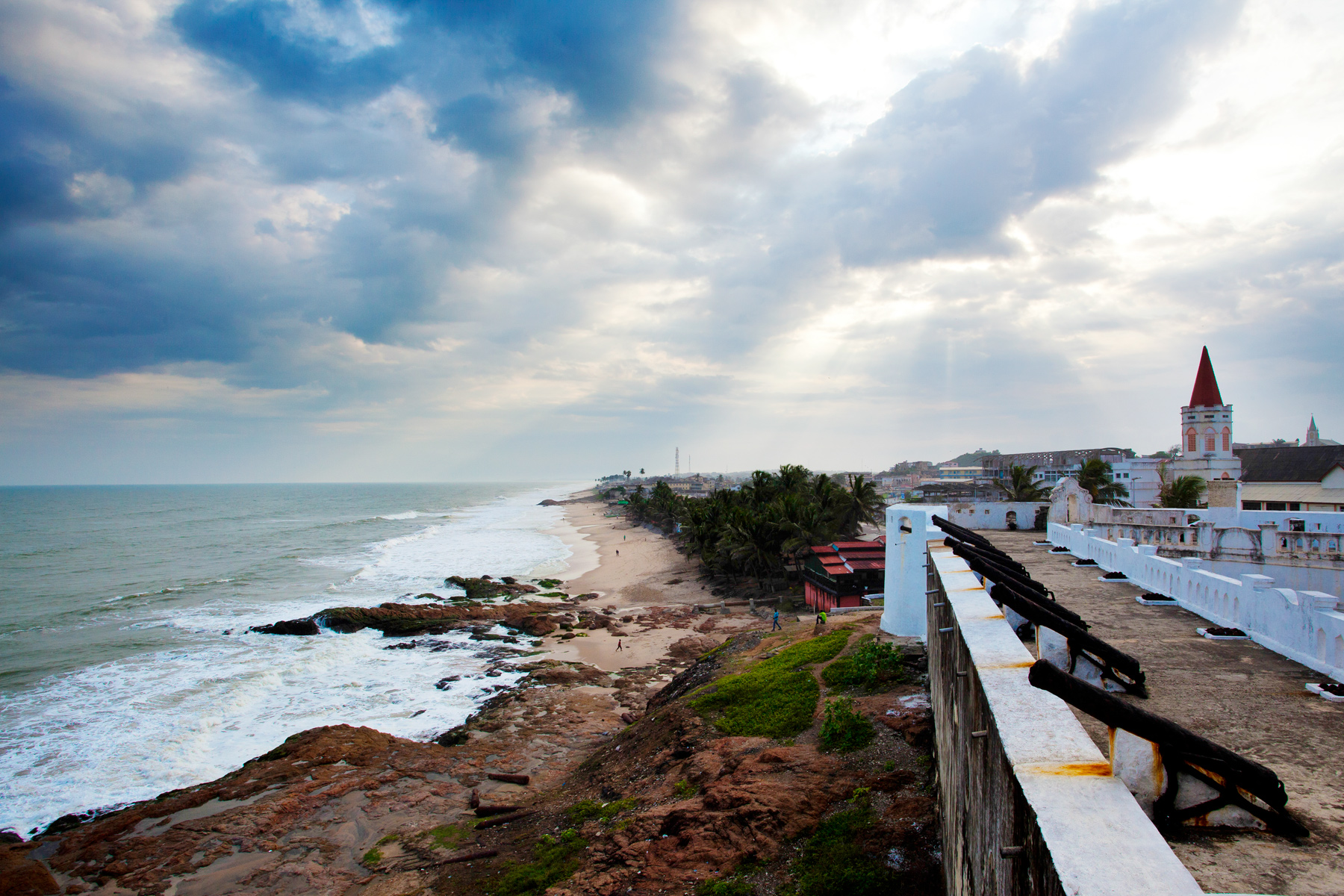 Coastal fort walls overlook a curving shoreline and town under a cloudy sky
