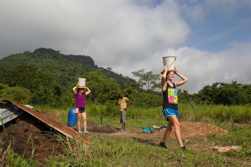 Two young women, dressed in casual attire, are carrying buckets on their heads while walking through a lush, rural area, with a man working in the background near a pile of earth, all set against a backdrop of green hills and cloudy skies.