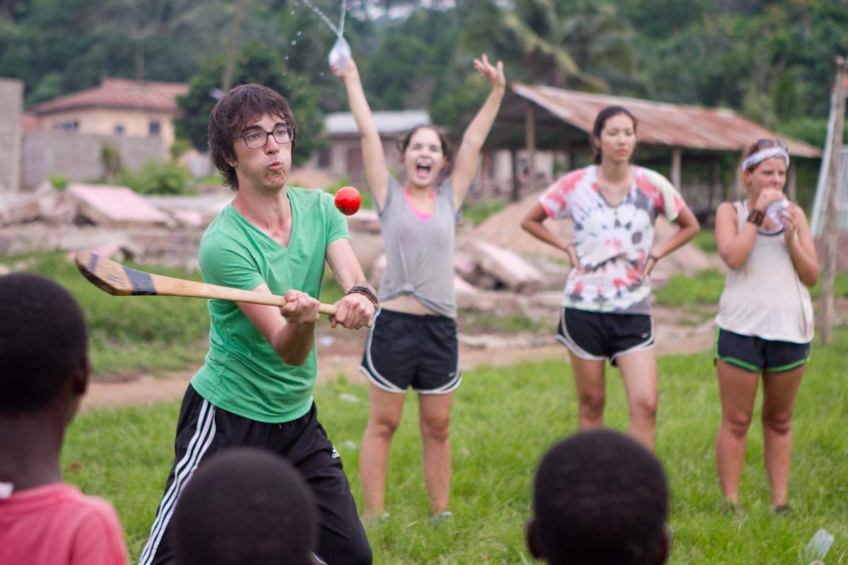A student swings a bat at a ball during an outdoor game as others cheer
