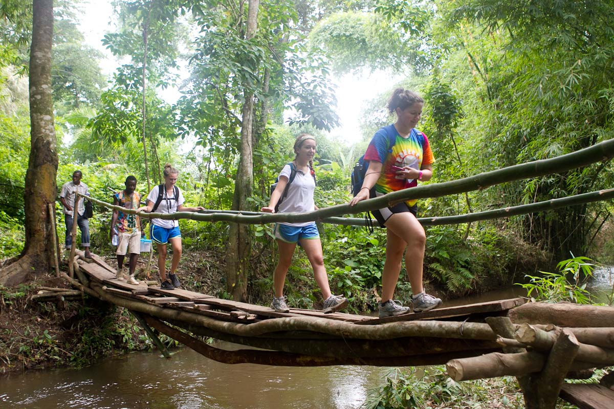 Students cross a wooden suspension bridge over a river in a forested area