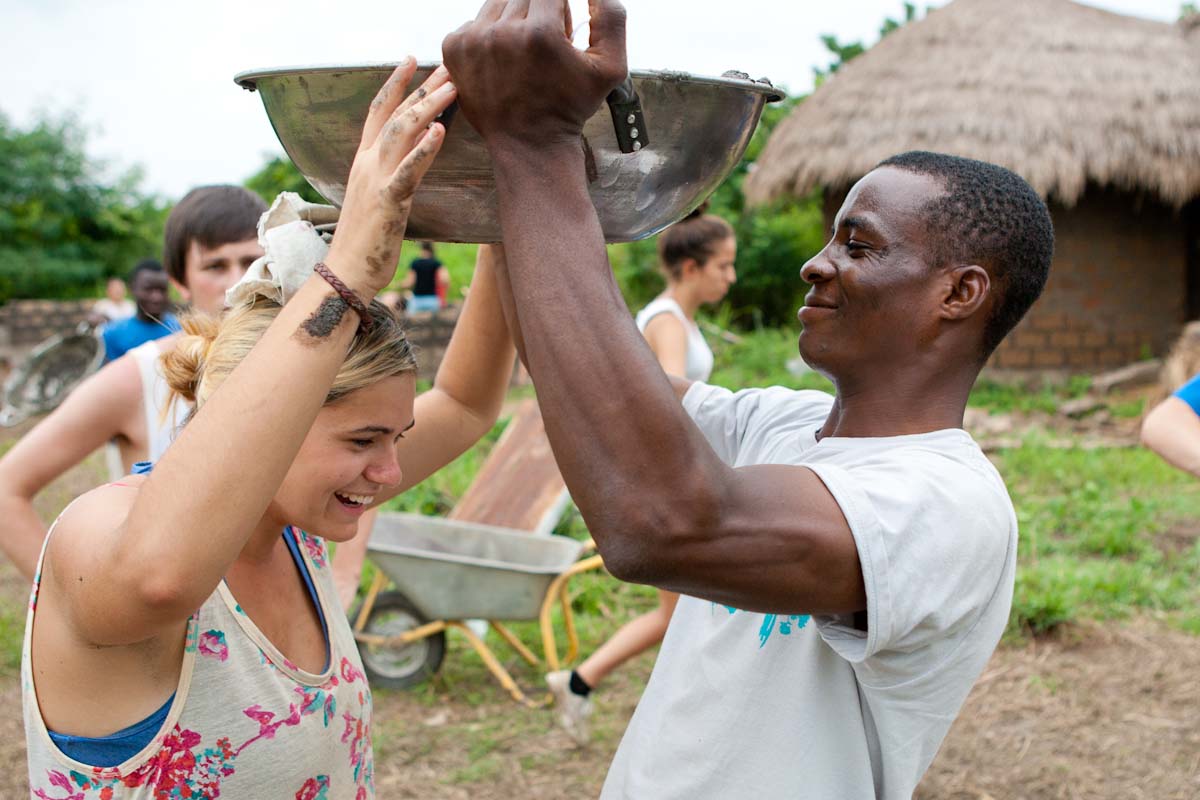 On this school trip, a local resident helps a student balance a metal basin on her head in a village setting