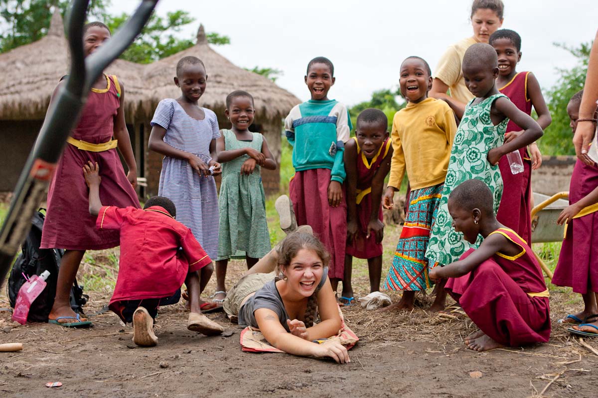 A student lies laughing on the ground surrounded by cheerful village children