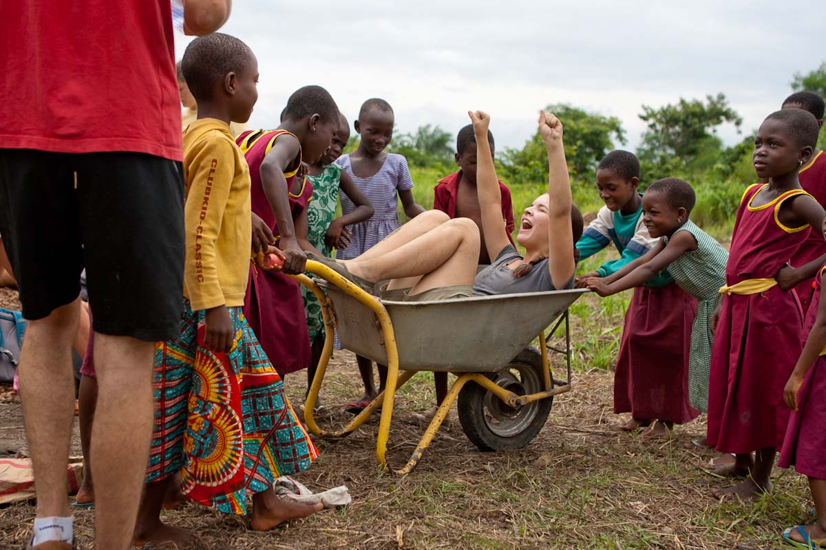 A student cheers while being pushed in a wheelbarrow as local children gather around laughing