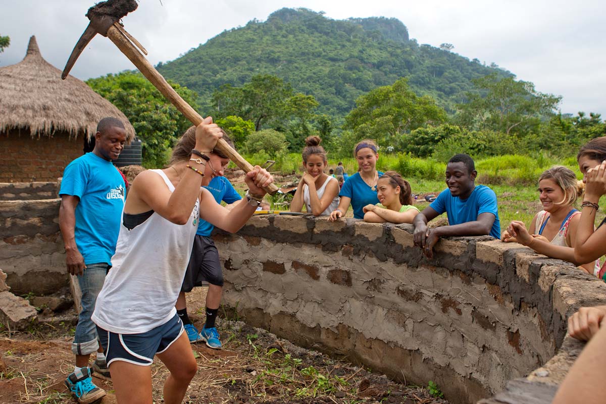 Students swing a pickaxe to help build a stone wall as others watch, with hills rising behind the village