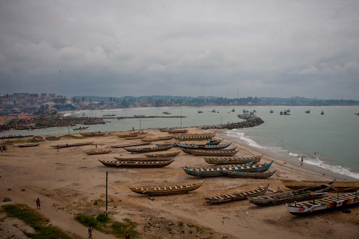 Wooden fishing boats line a sandy beach along the coast in Ghana under a cloudy sky