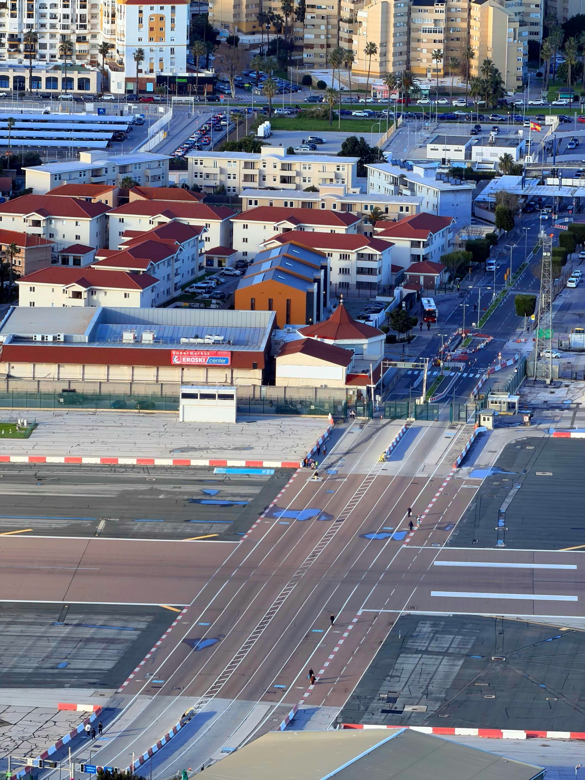 Seen from above, a runway's painted centerline crosses a road with barriers, low buildings and a few people on foot.
