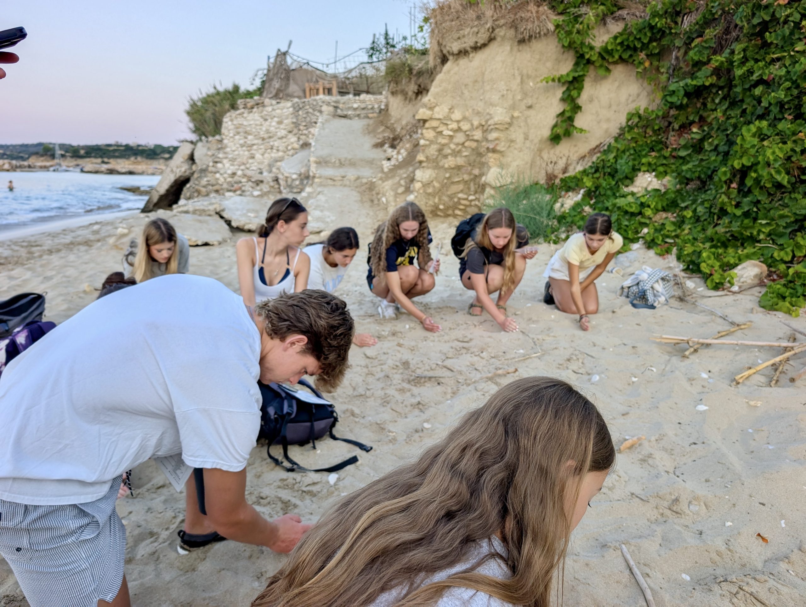 Students crouch on a beach searching the sand near a rocky hillside
