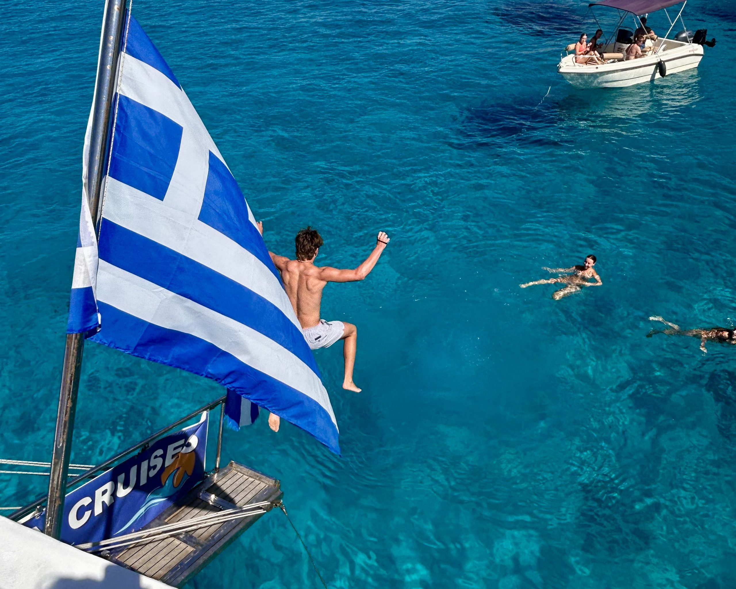 A student leaps off a boat past a Greek flag into clear blue water