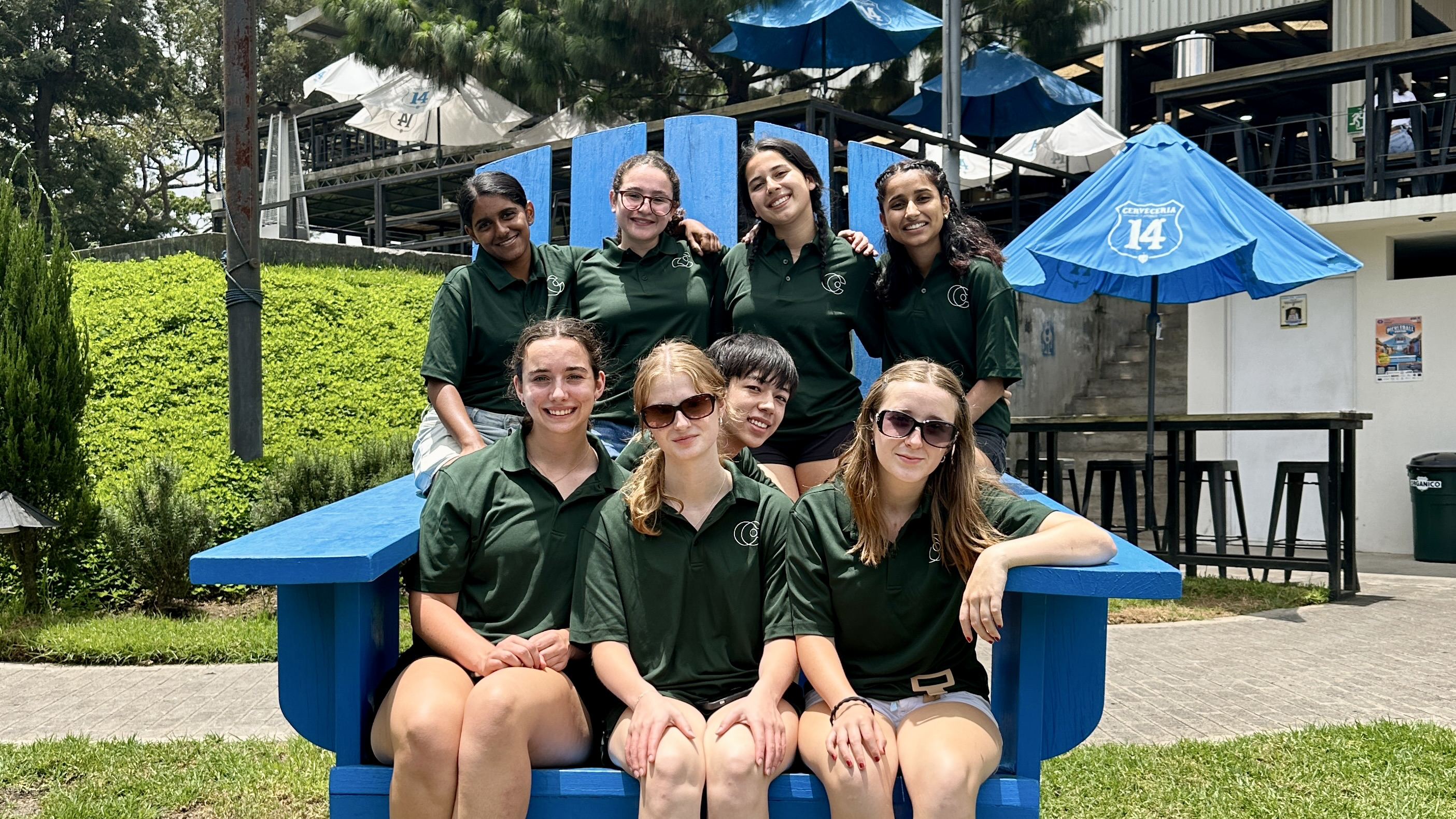 Students in matching green polos gather on an oversized blue bench outdoors
