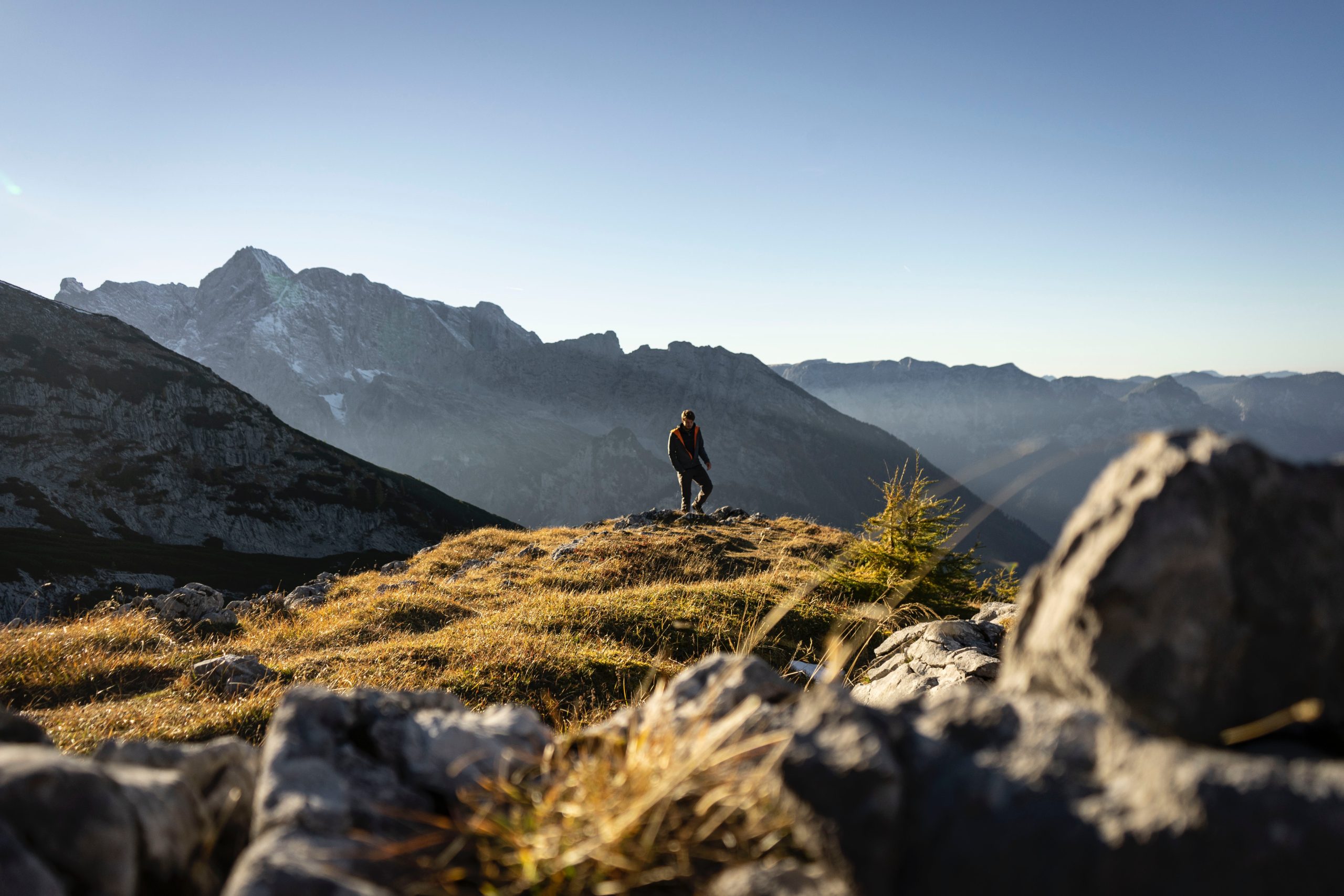 A student stands atop a grassy ridge overlooking layered mountain peaks