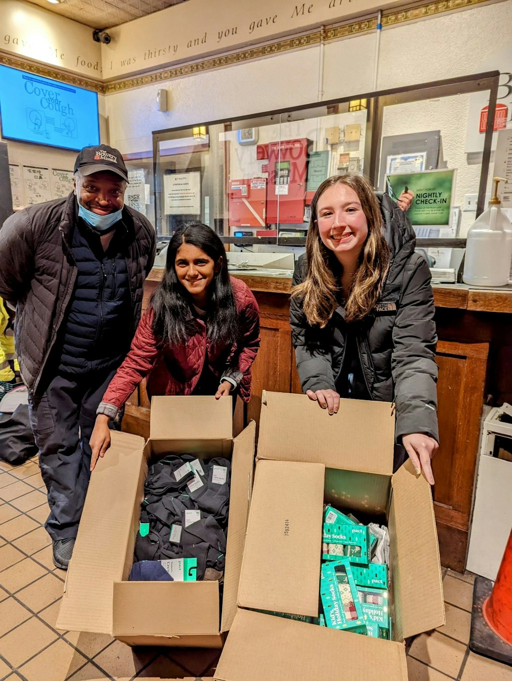 Two students and their mentor posing by a box of socks and underwear to give to the homeless. 