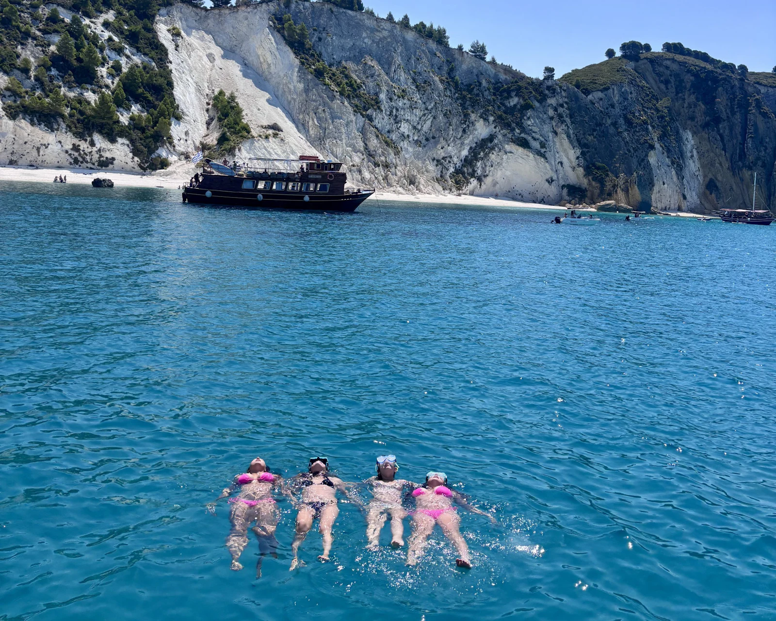 Four students float on their backs in deep blue water below white limestone cliffs in Greece, with a wooden excursion boat anchored behind