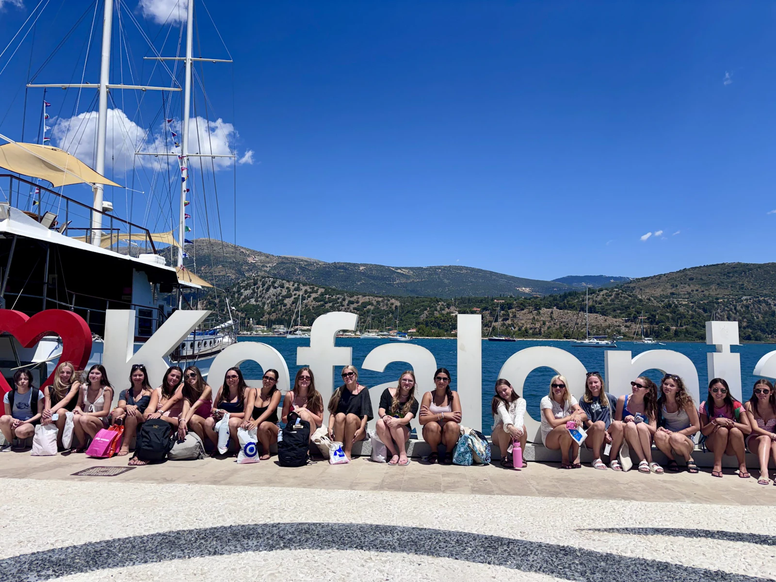 A Rustic Pathways group sits in a row along a large Kefalonia sign on the harbor front in Greece, with a sailing ship and green hills behind