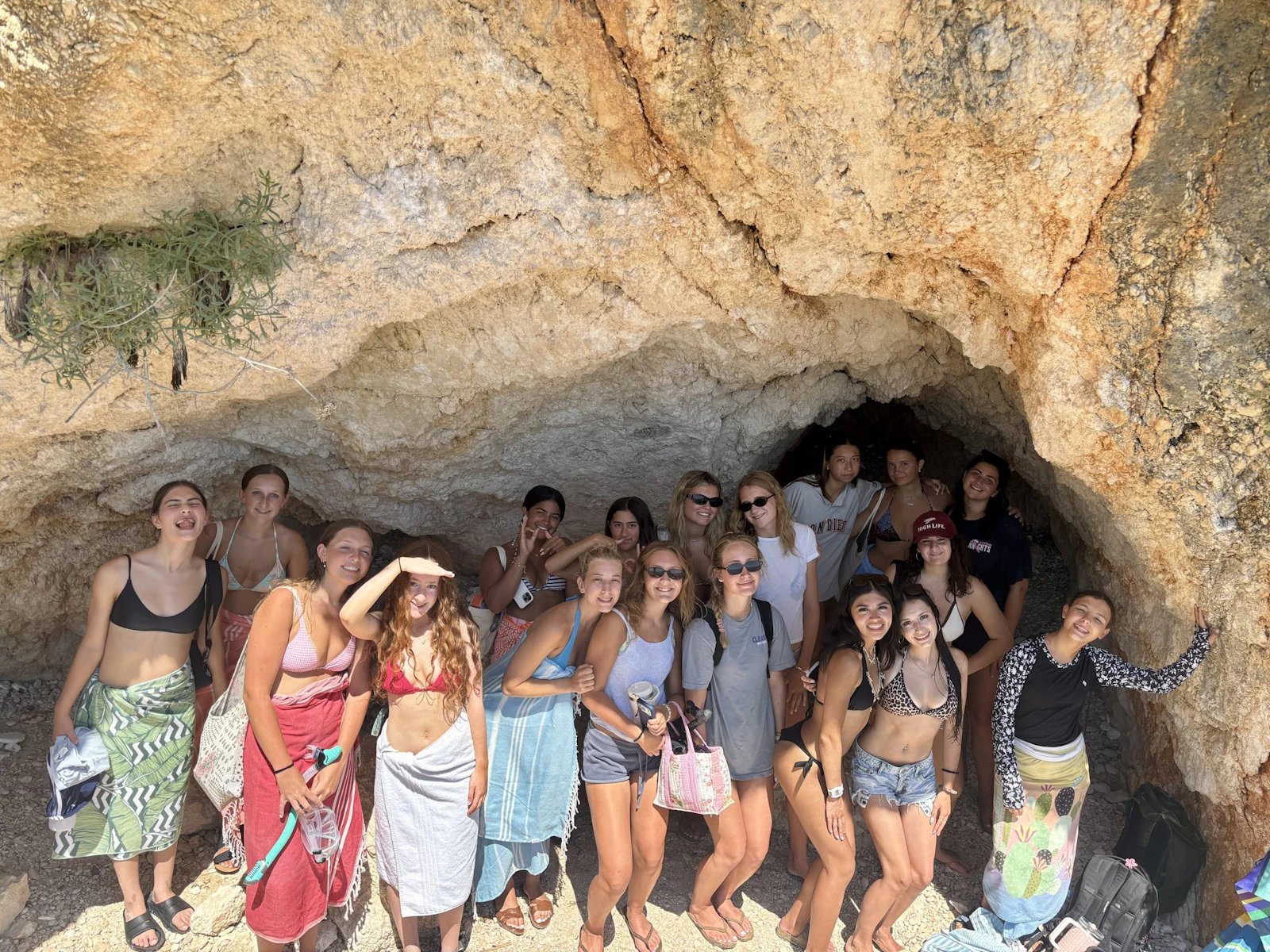 A Rustic Pathways group in swimsuits and sarongs stands beneath a rocky overhang at a sea cave in Greece