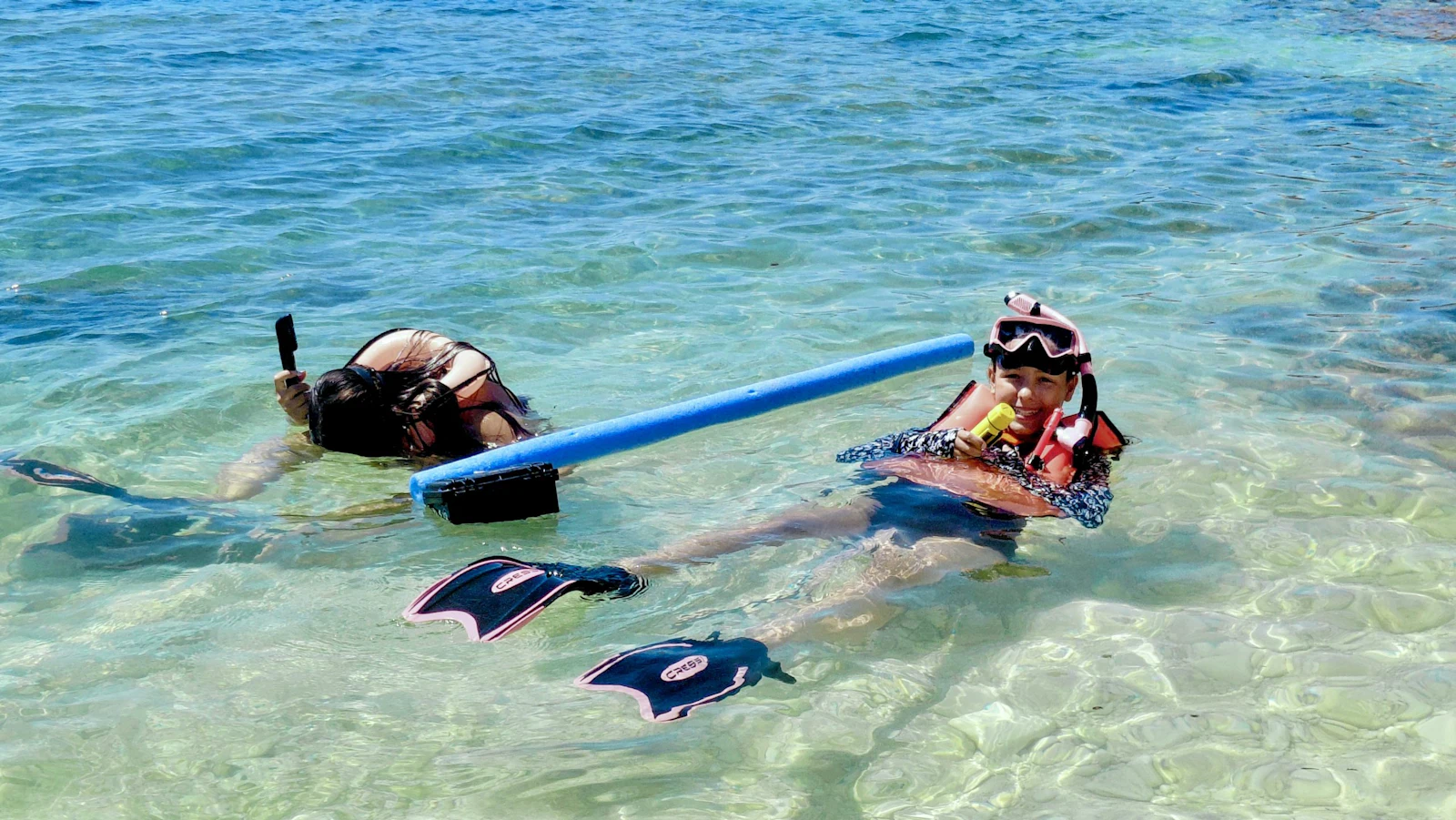 Two students in snorkels and fins float in clear shallow water in Greece, one holding a blue foam float