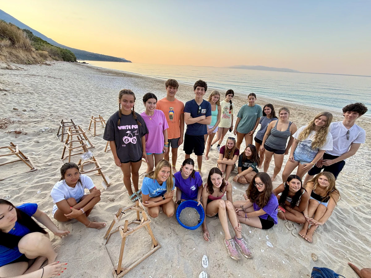 A Rustic Pathways group gathers at sunrise on a beach in Greece beside wooden nest-protection frames, one student holding a blue sieve