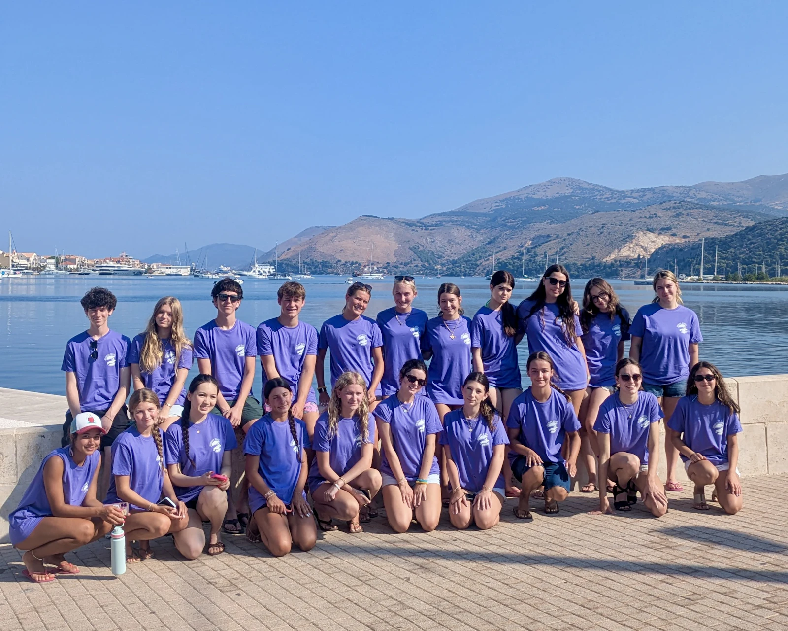 Students in matching purple shirts kneel and stand on a harbor walkway in Greece, with mountains across the bay