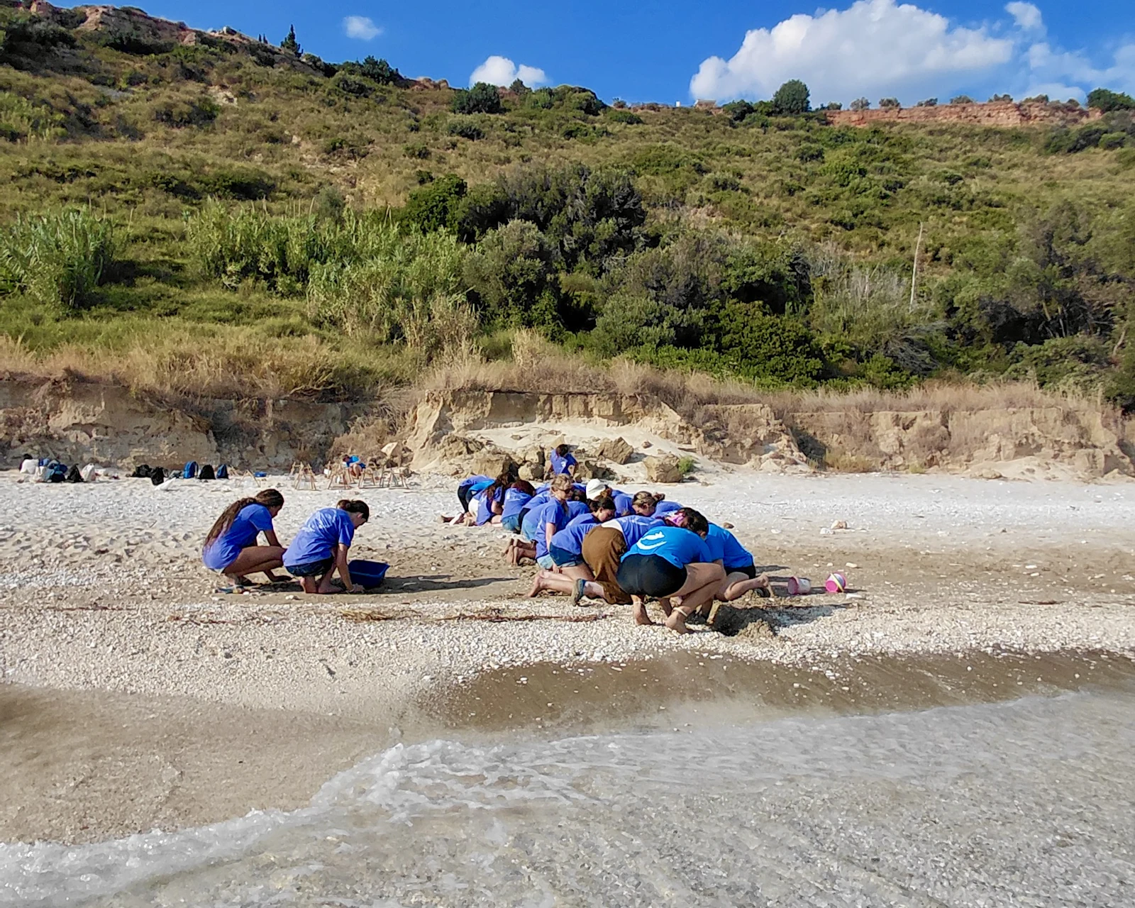 Students in blue shirts crouch at the edge of a pebbly beach in Greece, working on the sand below a scrubby hillside