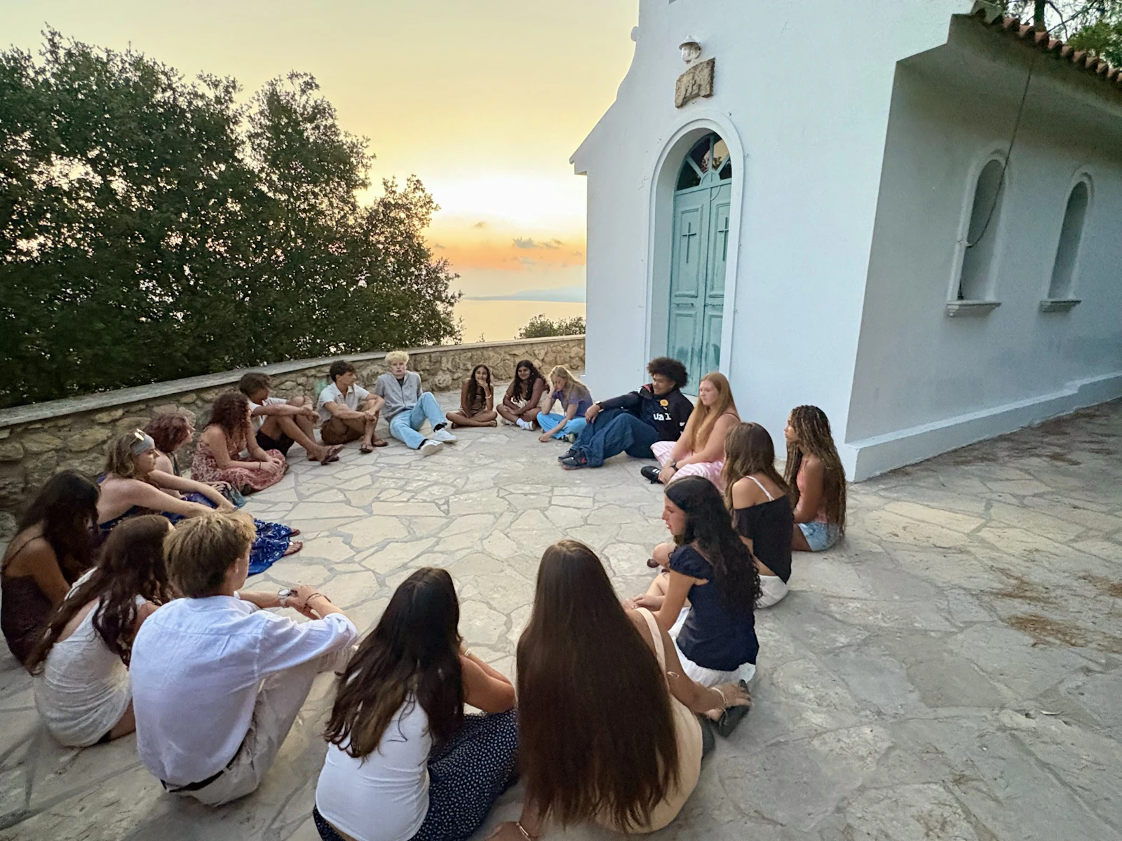 Students sit in a circle on a stone terrace beside a white chapel in Greece as the sun sets over the sea
