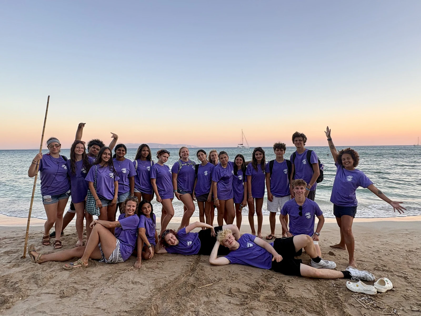 Students in matching purple shirts pose on a beach in Greece at sunset, one holding a tall wooden pole