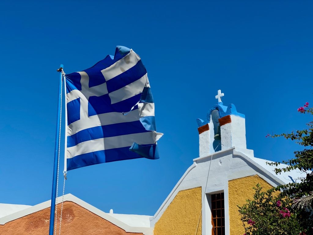The Greek flag waves in the wind against a striking blue sky and the top of a church building. Fun Facts About Greece
