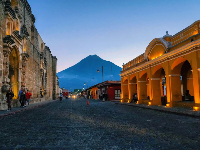 A cobbled street at dusk between a ruined stone church and a lit arcade, with a volcanic cone closing the view.