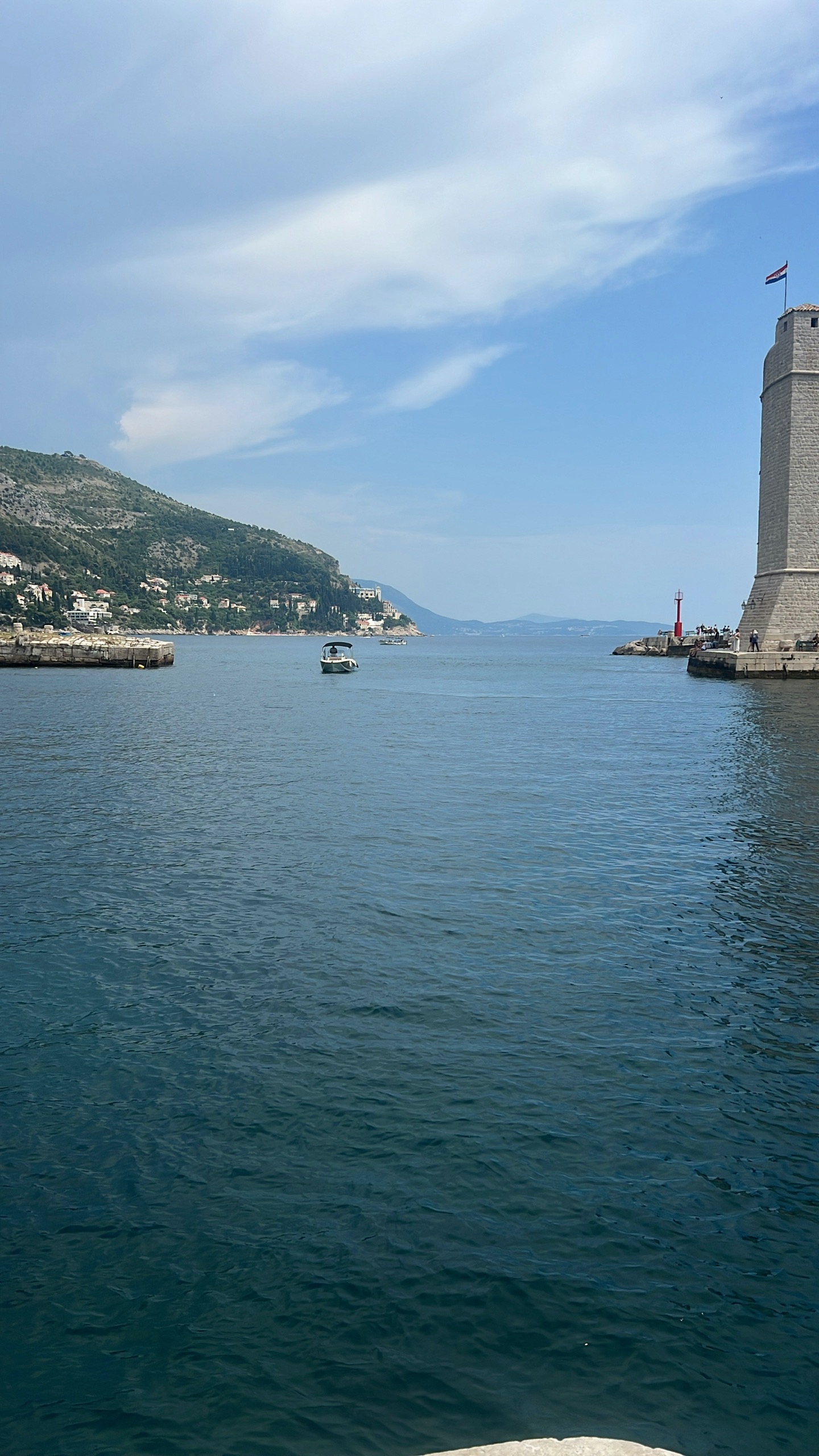 A small boat crosses the calm water at Dubrovnik's old harbor entrance, a stone fort and lighthouse beyond.
